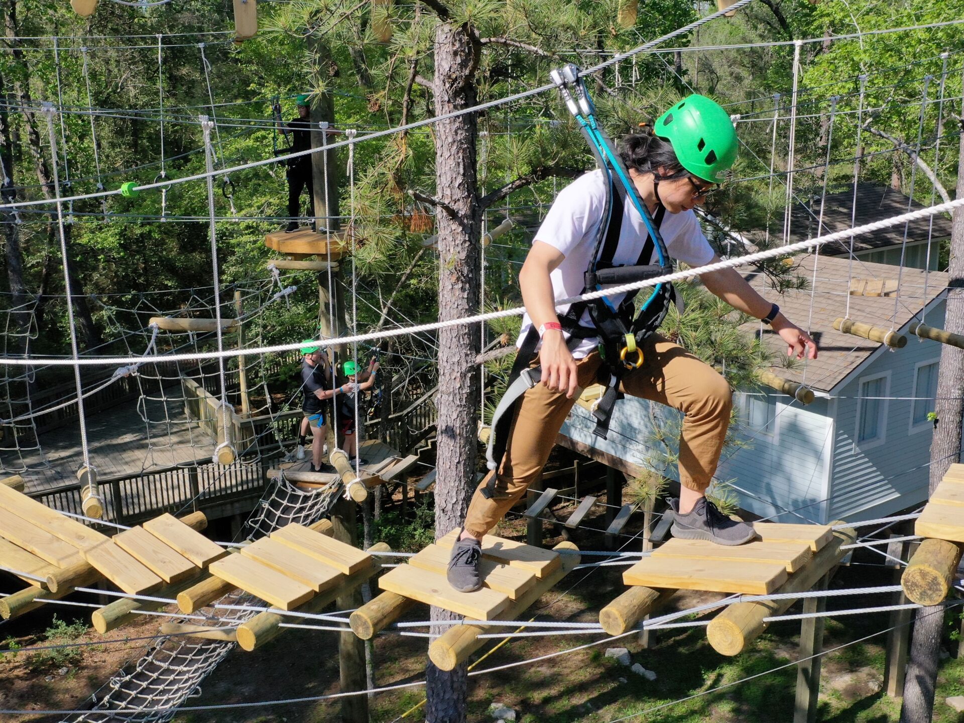 A group of people navigating the Texas TreeVentures course. They are wearing green helmets and harnesses for safety. The participants are balancing on wooden planks suspended by ropes, with one person in the foreground carefully stepping forward. Trees and additional ropes course elements are visible in the background, along with a building partially seen through the trees