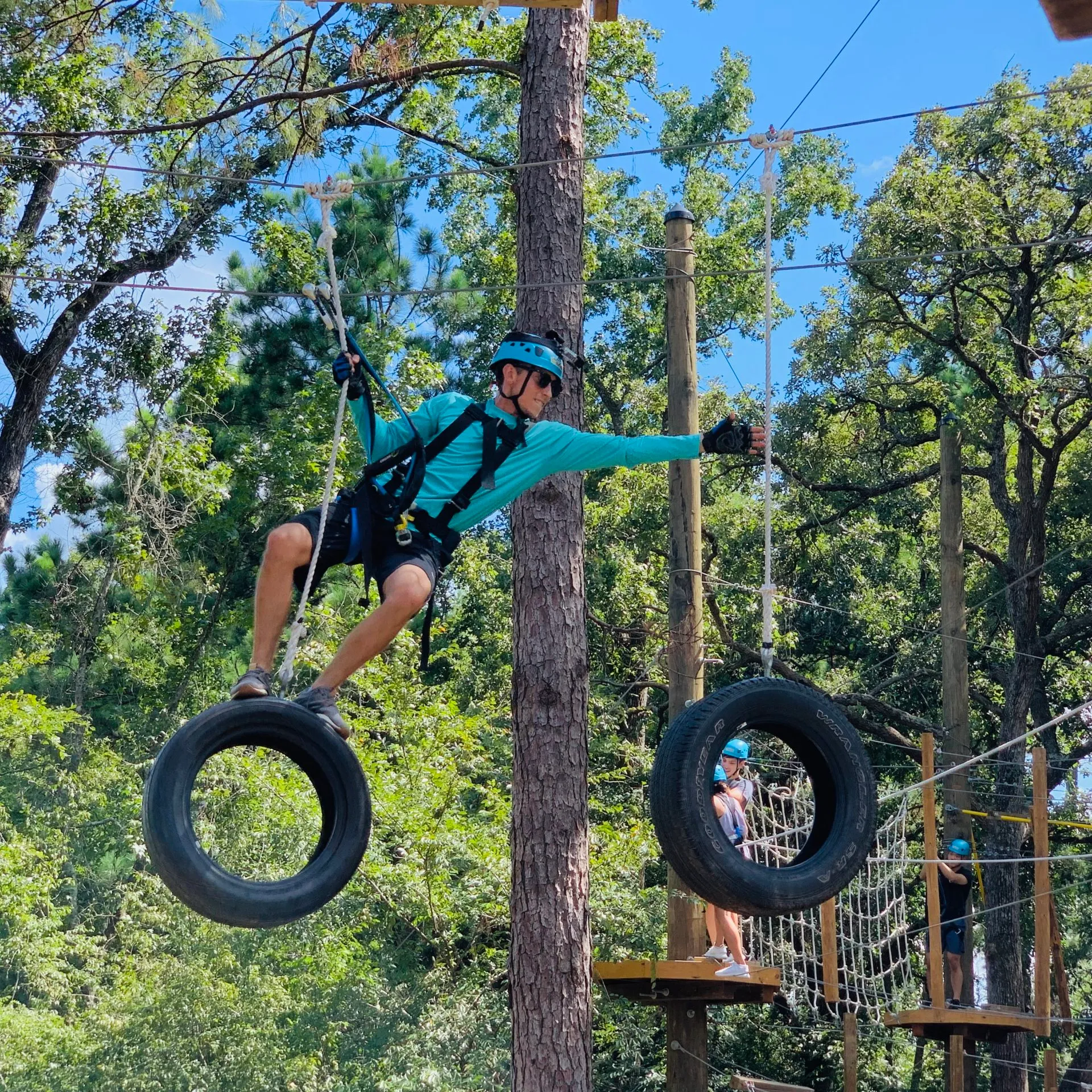 A person in a blue helmet and gloves, wearing a harness, navigates a ropes course by balancing on hanging tires. The participant is holding onto ropes attached to the tires, with one arm extended for balance. The background features a tall tree and other participants on the ropes course, along with various obstacles such as netting and wooden platforms. The sky is clear and blue, and the area is surrounded by lush greenery.