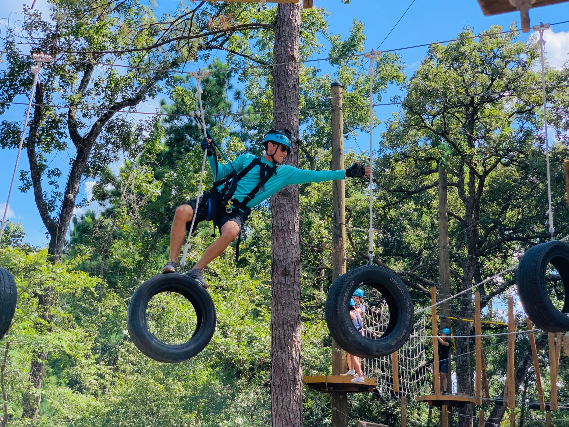 A person in a blue helmet and gloves, wearing a harness, navigates a ropes course by balancing on hanging tires. The participant is holding onto ropes attached to the tires, with one arm extended for balance. The background features a tall tree and other participants on the ropes course, along with various obstacles such as netting and wooden platforms. The sky is clear and blue, and the area is surrounded by lush greenery.