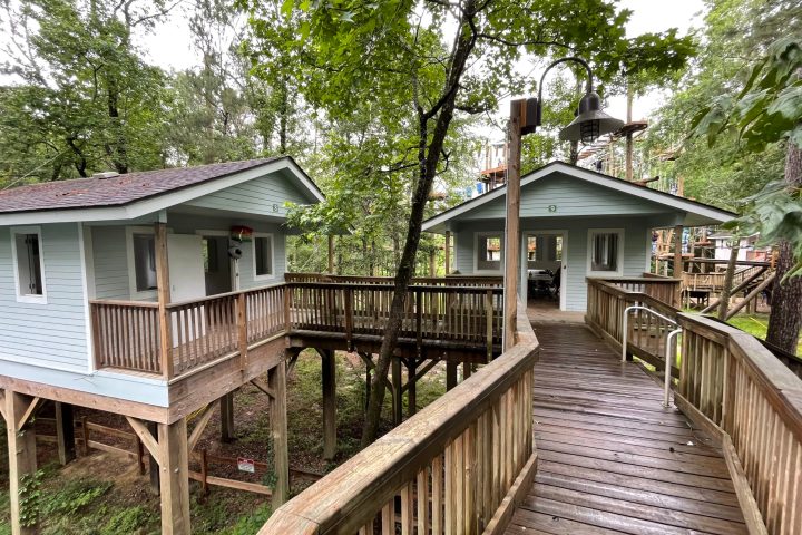 A view of two elevated cabins connected by a wooden walkway in a forested area. The cabins, painted light blue with white trim, have porches with railings and are supported by wooden stilts. The walkway leading to the cabins is also made of wood and has railings for safety. The surrounding area is lush with trees and greenery, and part of a ropes course structure is visible in the background.