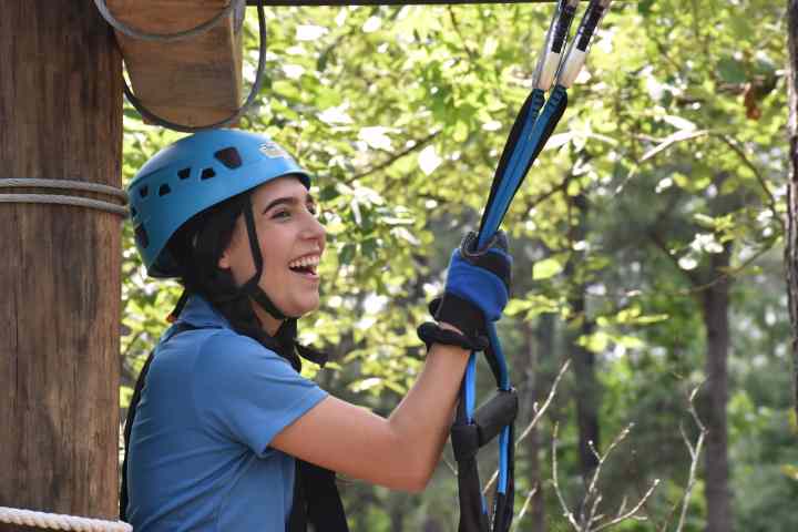 A smiling woman wearing a blue helmet and matching gloves is standing next to a wooden pole on a ropes course. She is holding onto a safety harness with both hands, preparing to engage in the course. The background features lush green foliage, indicating a forested area. The woman appears excited and happy, ready to participate in the adventure activity.