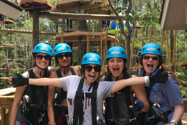 A group of five women are posing together, smiling and laughing, while wearing blue helmets and harnesses on a ropes course. They are standing on a wooden platform with various climbing and adventure elements visible in the background, such as ropes, wooden platforms, and trees. The women are clearly excited and ready to take on the adventure course, with one woman in the center extending her arms wide in a gesture of enthusiasm.