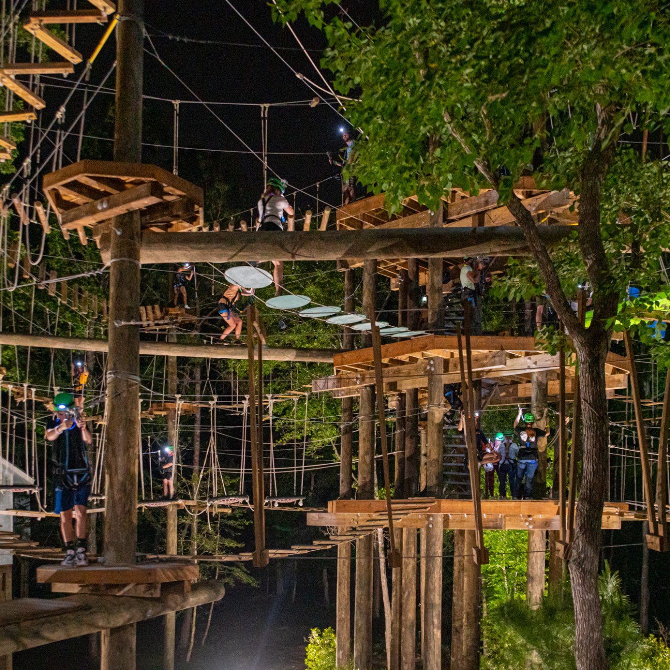 A nighttime view of a multi-level ropes course filled with participants. The structure is made of wooden platforms, ropes, and various obstacles, including hanging steps, planks, and circular platforms. Participants are equipped with helmets and harnesses, navigating the course with headlamps and artificial lighting. The background is dark, with trees and the ropes course structure illuminated by the lights.