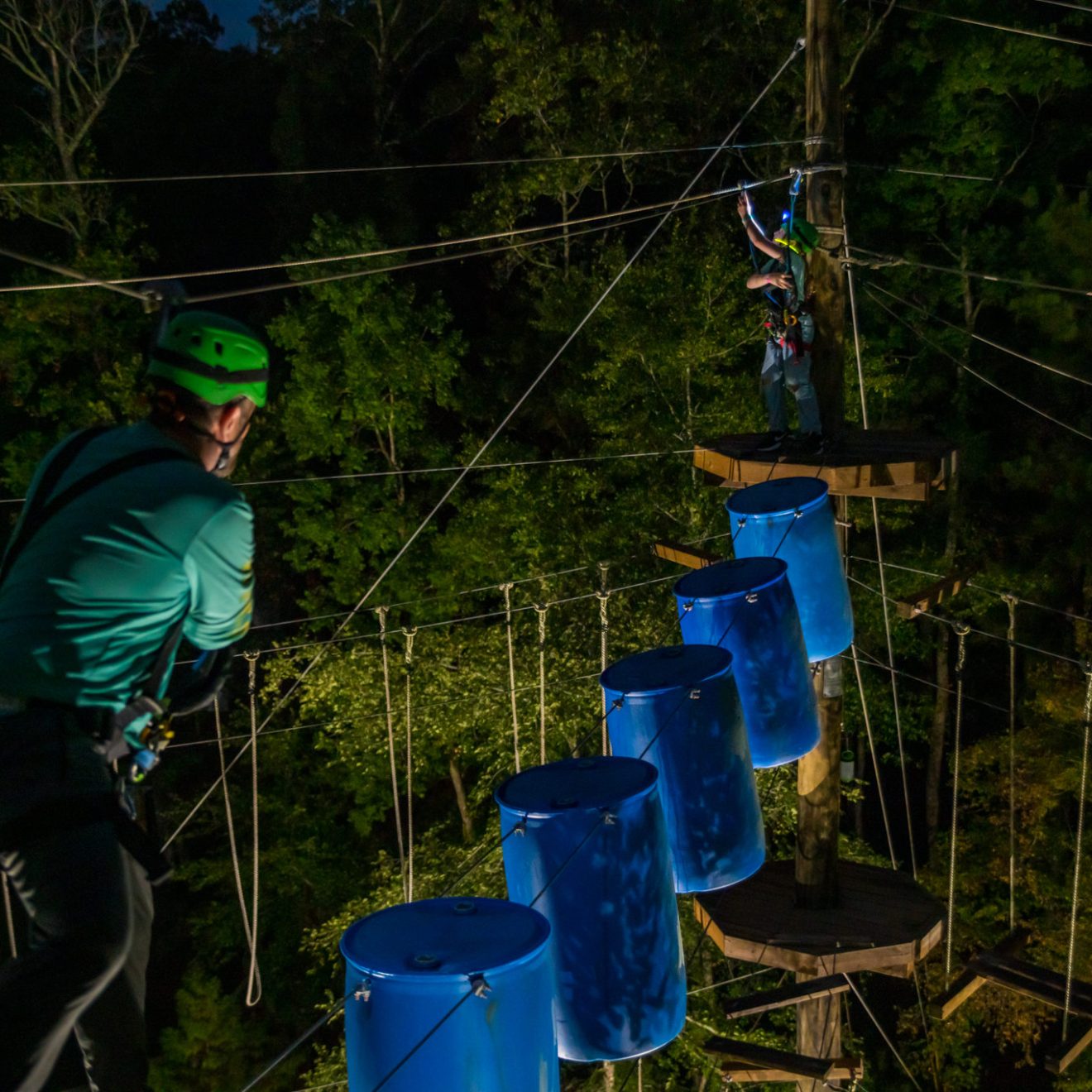Two people are participating in a nighttime ropes course activity. One person, closer to the camera, is wearing a green helmet and a harness while walking on a wooden plank. The second person is in the background, standing on a platform and gesturing, also equipped with safety gear. Blue barrels are suspended along the course, and the area is lit by artificial lighting, highlighting the trees and ropes.