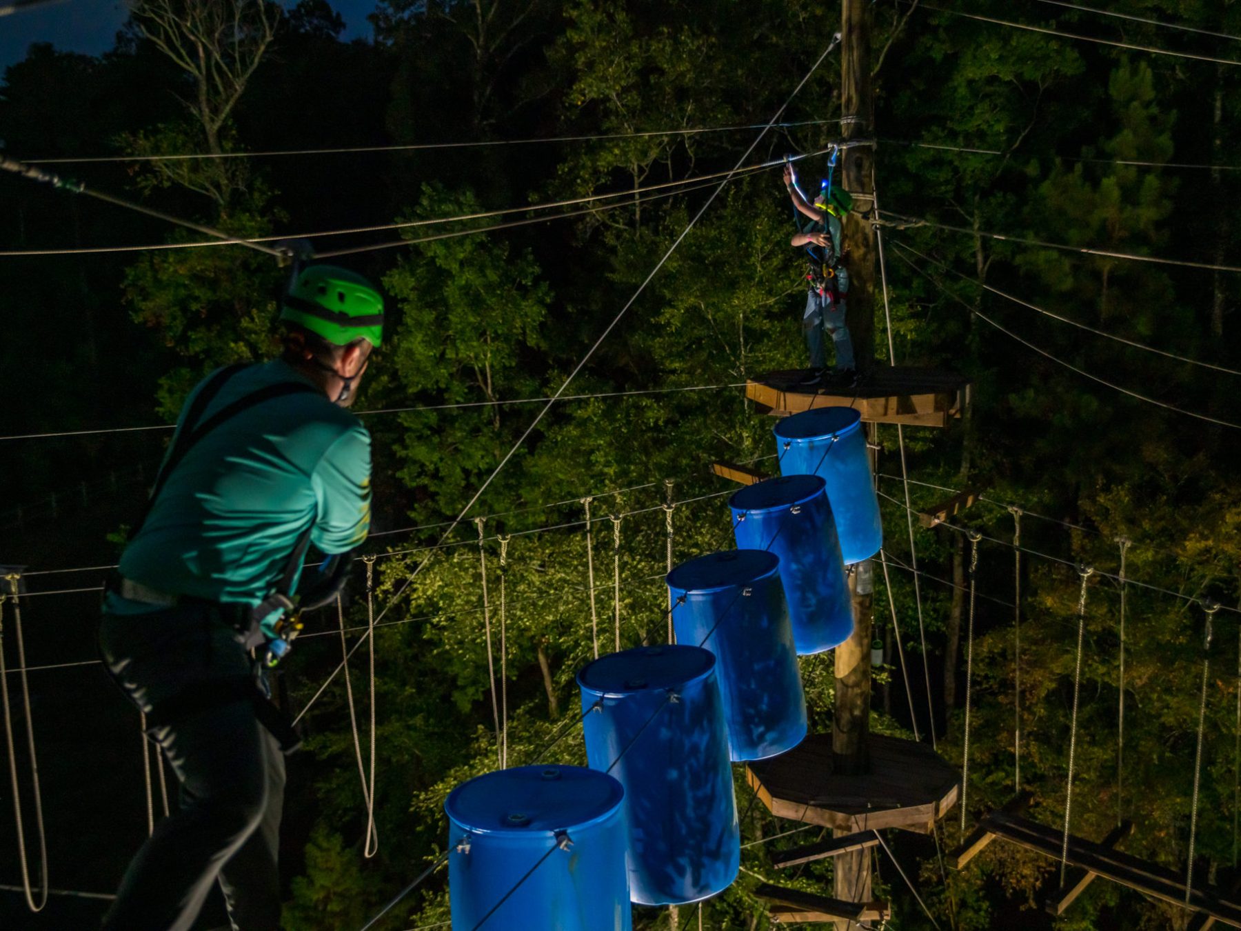 Two people are participating in a nighttime ropes course activity. One person, closer to the camera, is wearing a green helmet and a harness while walking on a wooden plank. The second person is in the background, standing on a platform and gesturing, also equipped with safety gear. Blue barrels are suspended along the course, and the area is lit by artificial lighting, highlighting the trees and ropes.