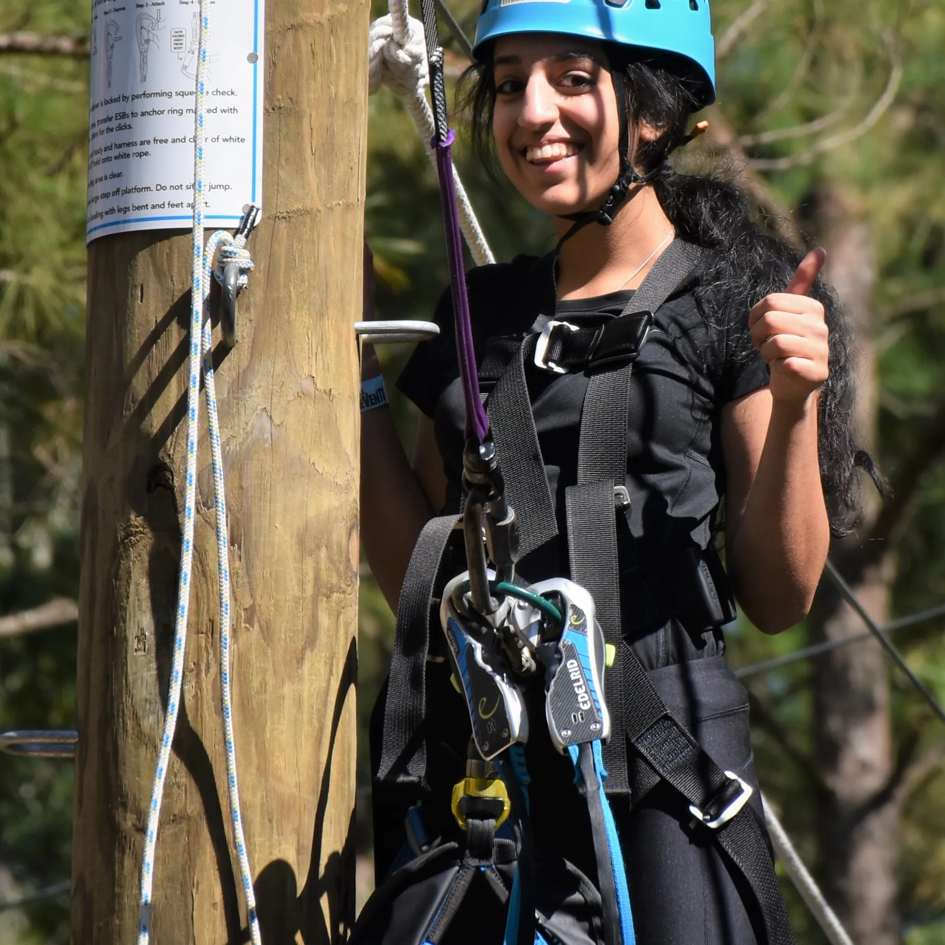 A person wearing a blue helmet and a safety harness smiles and gives a thumbs-up while standing on a platform of an aerial adventure course. The individual is next to a wooden post with instructional signage, and the background shows ropes and trees.