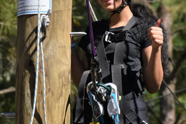 A person wearing a blue helmet and a safety harness smiles and gives a thumbs-up while standing on a platform of an aerial adventure course. The individual is next to a wooden post with instructional signage, and the background shows ropes and trees.