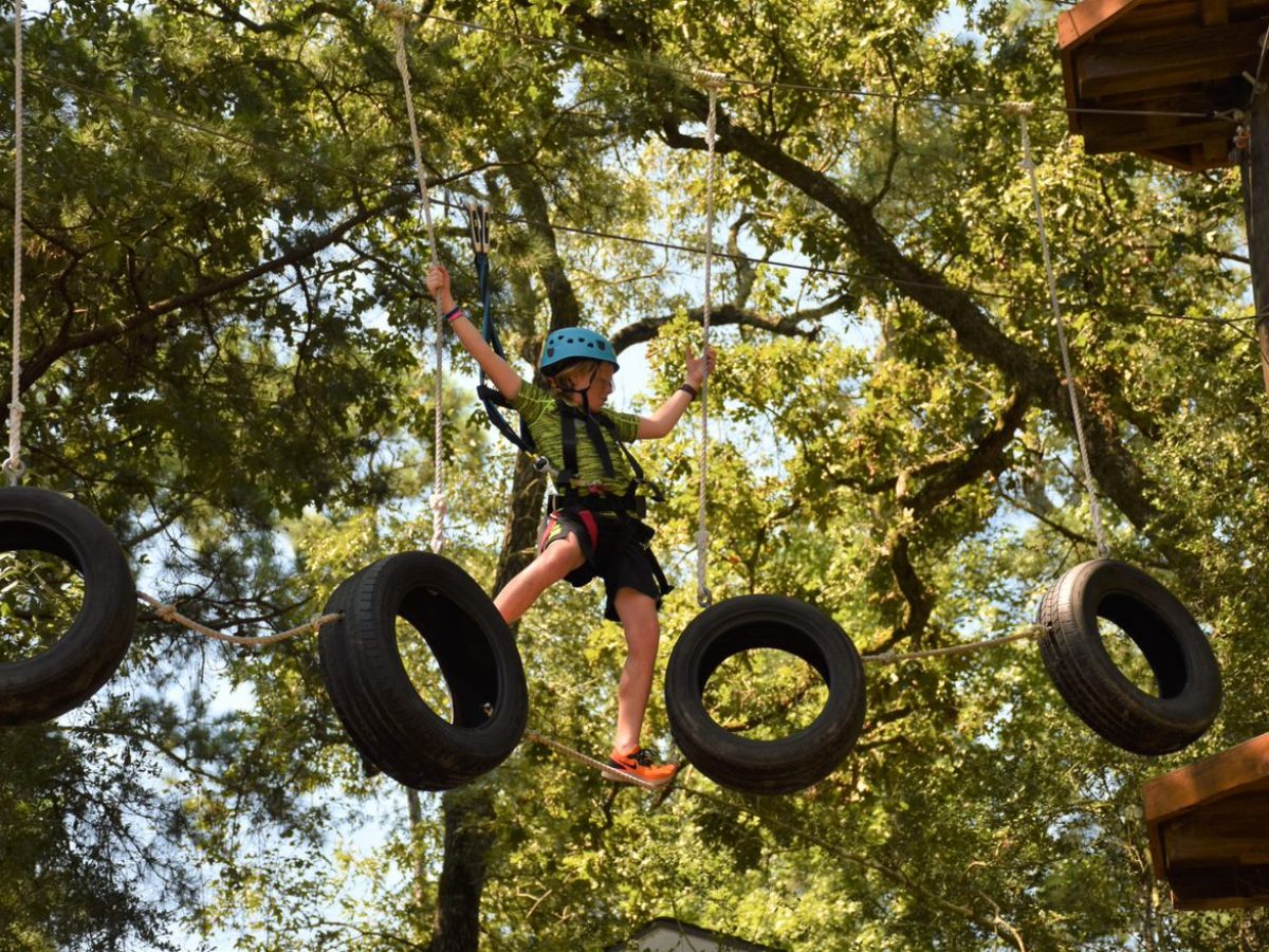 A person wearing a blue helmet and safety harness navigates a suspended obstacle course made of hanging tires. They are balancing on one of the tires with one leg extended to another tire, holding onto the safety ropes for support. The background features a lush green canopy of trees, suggesting a forested environment. The person appears focused and engaged in the activity.