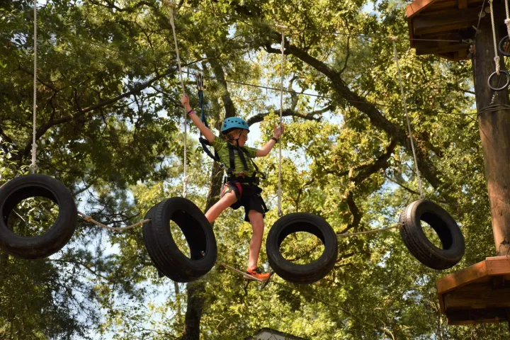 A person wearing a blue helmet and safety harness navigates a suspended obstacle course made of hanging tires. They are balancing on one of the tires with one leg extended to another tire, holding onto the safety ropes for support. The background features a lush green canopy of trees, suggesting a forested environment. The person appears focused and engaged in the activity.