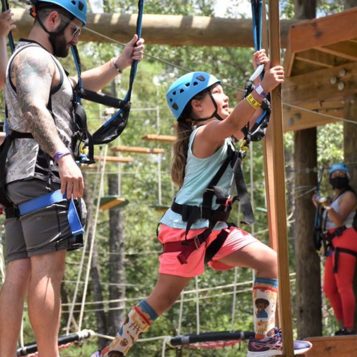 A young girl wearing a blue helmet and safety harness is climbing a wooden structure at an adventure park. She is accompanied by an adult, who is also wearing a blue helmet and safety harness. The girl is wearing colorful socks and athletic shoes. The background shows other participants engaged in the adventure course activities among trees and wooden platforms.