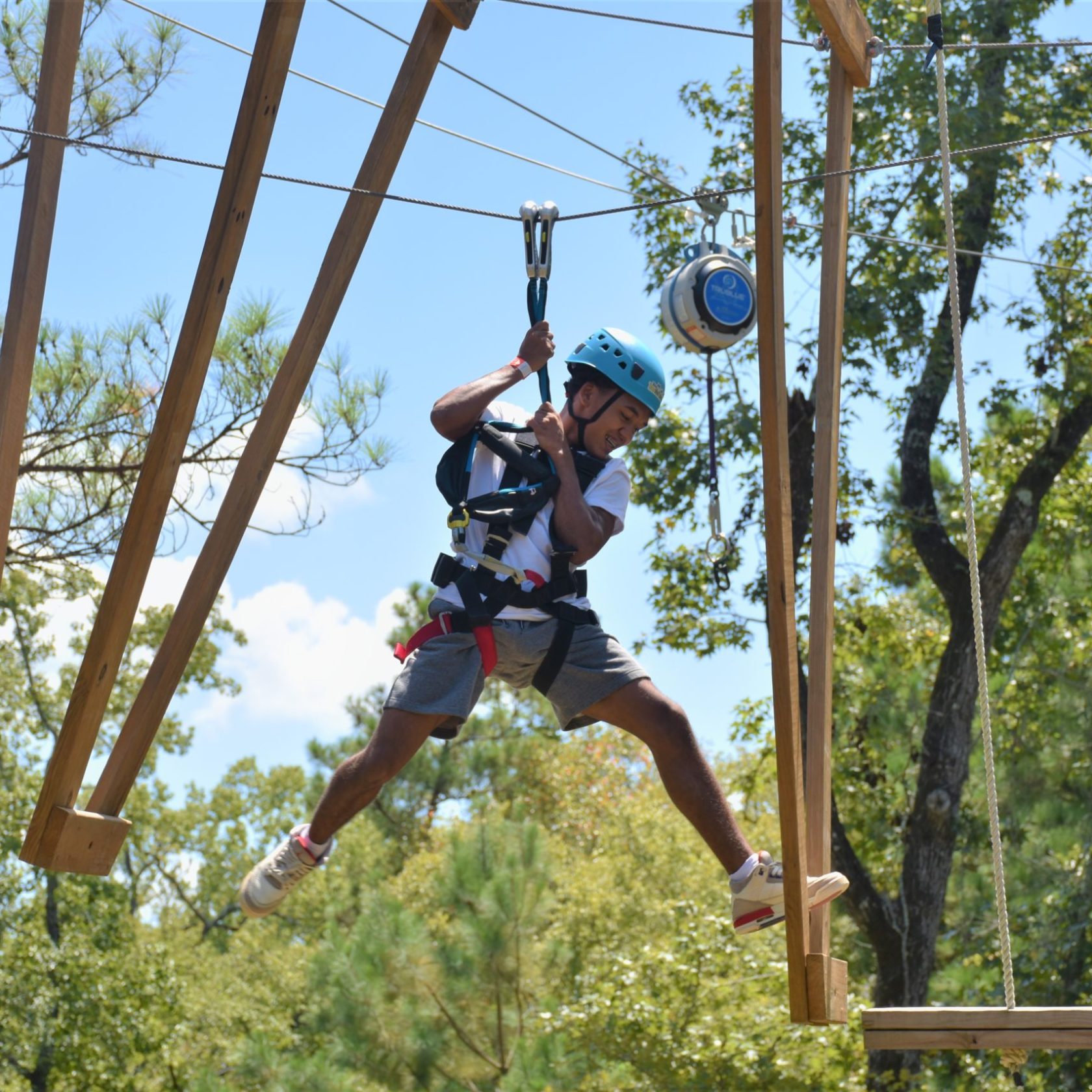 A person wearing a blue helmet and safety harness is engaged in an outdoor ropes course. They are holding onto their belay system while leaping toward a suspended wooden platform. The individual appears to be mid-movement, with one foot off the platform and an arm bent for balance. The background features a clear blue sky and tall green trees, adding to the adventurous setting.