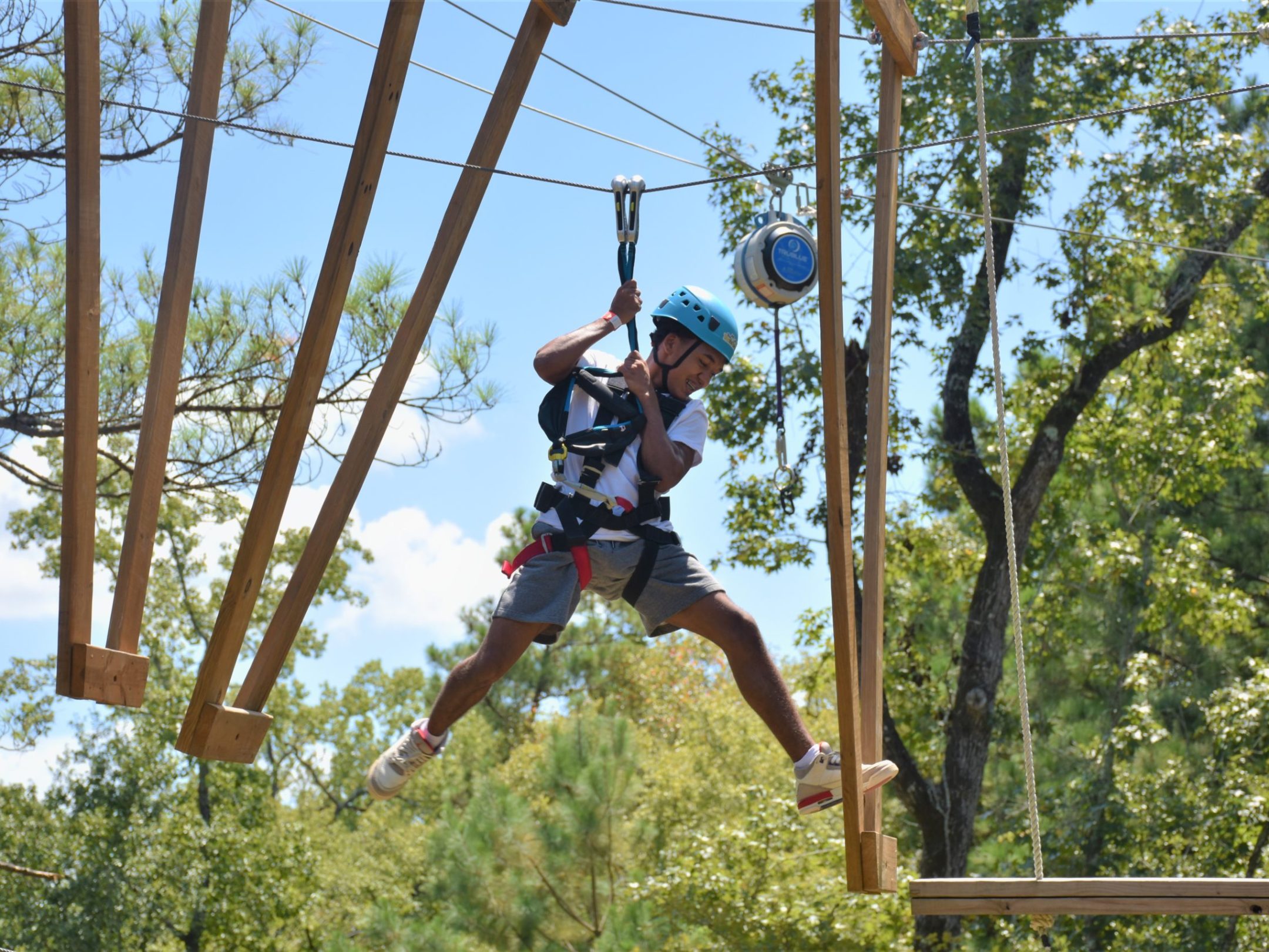 A person wearing a blue helmet and safety harness is engaged in an outdoor ropes course. They are holding onto their belay system while leaping toward a suspended wooden platform. The individual appears to be mid-movement, with one foot off the platform and an arm bent for balance. The background features a clear blue sky and tall green trees, adding to the adventurous setting.