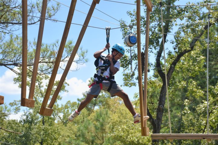 A person wearing a blue helmet and safety harness is engaged in an outdoor ropes course. They are holding onto their belay system while leaping toward a suspended wooden platform. The individual appears to be mid-movement, with one foot off the platform and an arm bent for balance. The background features a clear blue sky and tall green trees, adding to the adventurous setting.
