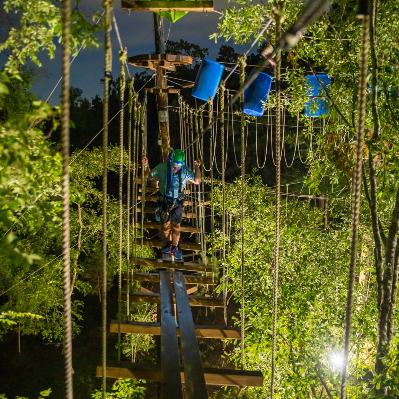 A nighttime view of an adventure ropes course illuminated by artificial lights. The course is surrounded by dense greenery, with a participant navigating a rope bridge. The participant is wearing a harness, helmet, and headlamp, adding to the safety and visibility. Blue barrels and various obstacles can be seen in the background, creating a challenging and adventurous atmosphere.