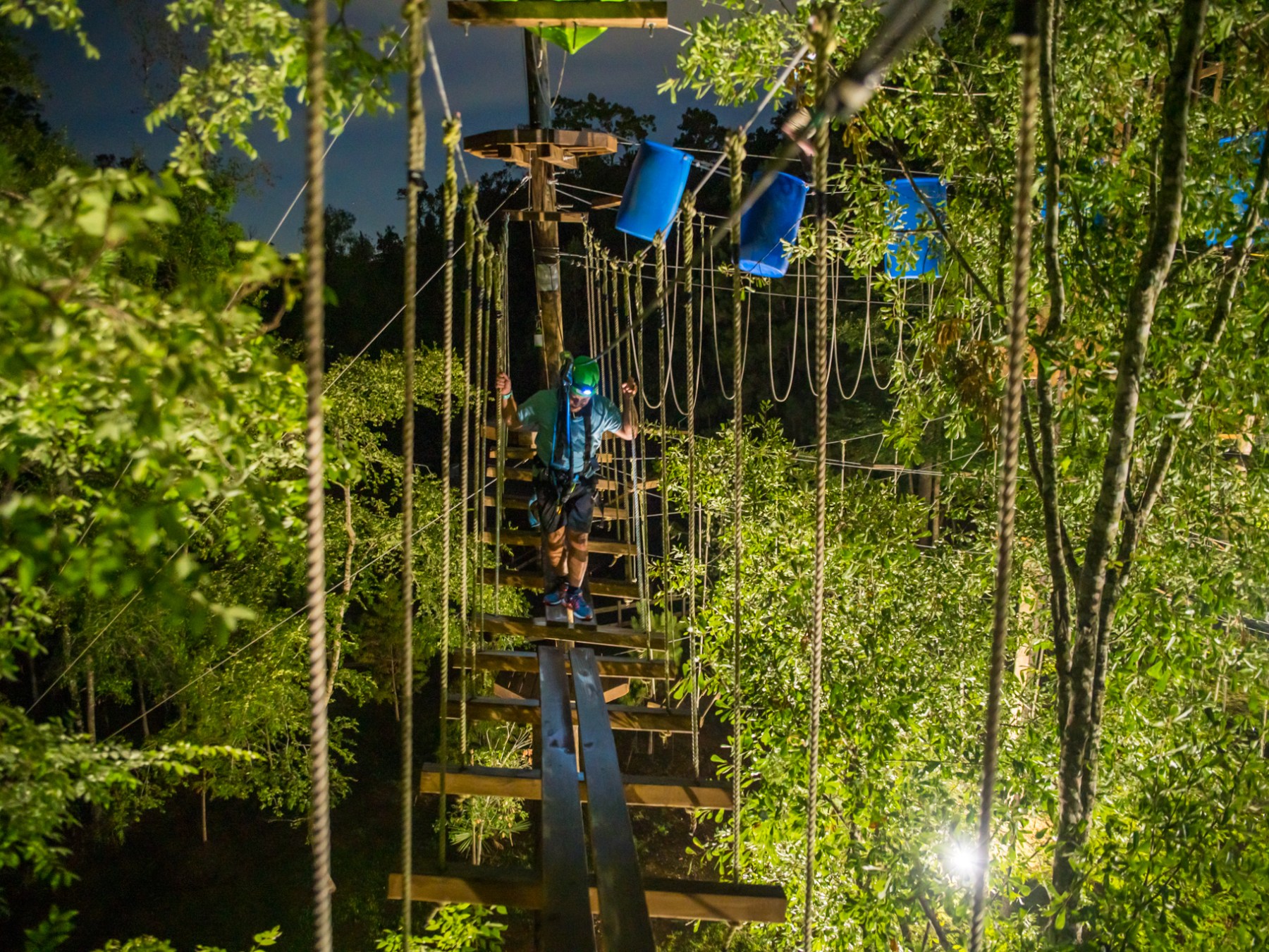 A nighttime view of an adventure ropes course illuminated by artificial lights. The course is surrounded by dense greenery, with a participant navigating a rope bridge. The participant is wearing a harness, helmet, and headlamp, adding to the safety and visibility. Blue barrels and various obstacles can be seen in the background, creating a challenging and adventurous atmosphere.