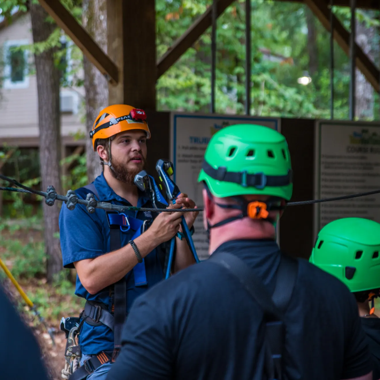 A group of people in green helmets and harnesses listen attentively to an instructor, who is demonstrating safety procedures on a ropes course. The instructor, wearing an orange helmet and a blue shirt, holds safety gear while speaking. The background features a wooded area with trees and informational signs about the course rules.