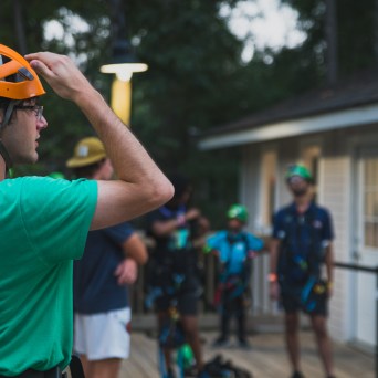 A man wearing an orange helmet and green shirt adjusts his helmet while standing on a wooden deck. In the background, there are other people, including adults and children, also wearing helmets and harnesses, preparing for the ropes course. The scene takes place near a building under an outdoor light, and the background is slightly blurred, focusing on the man in the foreground.