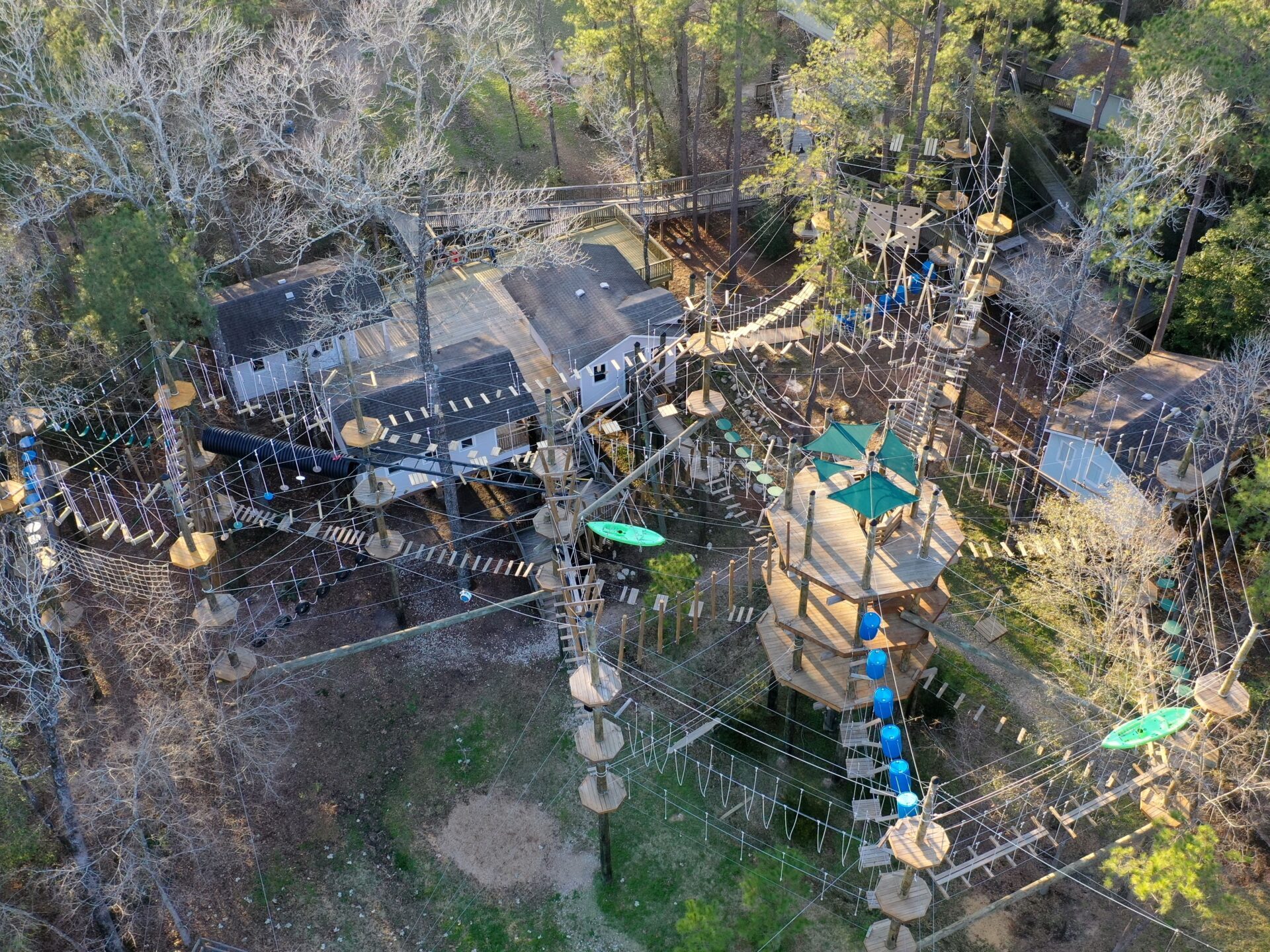 An aerial view of a high ropes course surrounded by trees. The course features various obstacles, including platforms, ropes, and suspended bridges. There are several levels to the course, with participants wearing helmets and safety harnesses. A green kayak is suspended as part of the course. The area is well-structured, with a combination of wooden platforms and ropes, offering a challenging adventure experience. Buildings and pathways are visible in the background, blending with the forest setting.