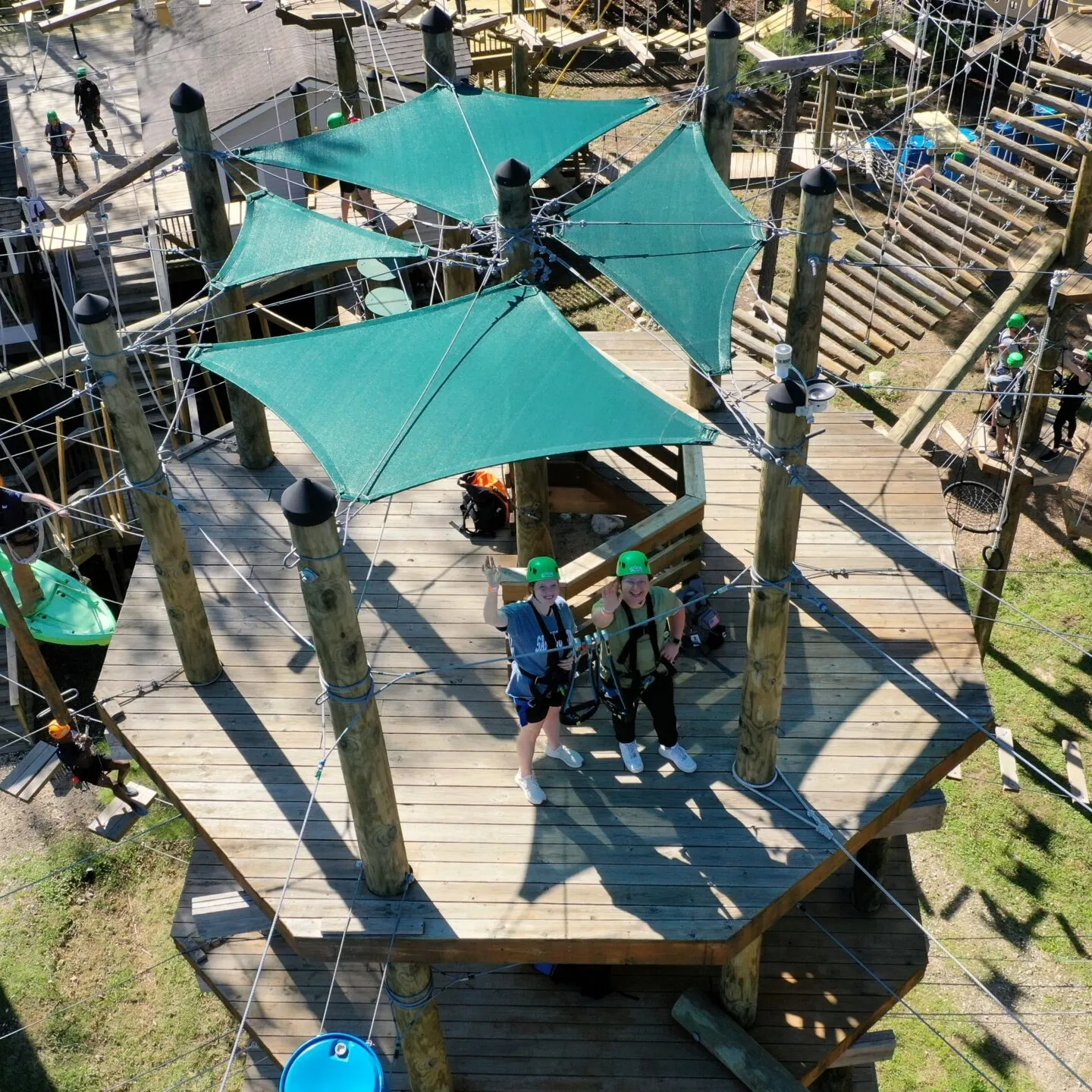 An aerial view of a section of an outdoor high ropes course. The course includes a large wooden platform with green shade canopies and several participants wearing helmets and safety harnesses. The platform is part of a complex network of ropes, obstacles, and wooden structures. Other participants and various course elements are visible in the background, set among trees and foliage, indicating an outdoor environment.