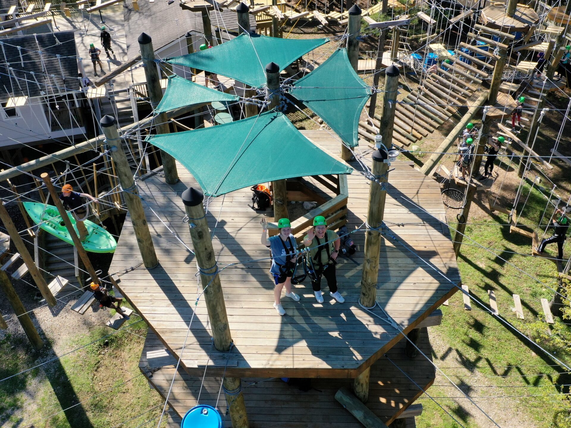 An aerial view of a section of an outdoor high ropes course. The course includes a large wooden platform with green shade canopies and several participants wearing helmets and safety harnesses. The platform is part of a complex network of ropes, obstacles, and wooden structures. Other participants and various course elements are visible in the background, set among trees and foliage, indicating an outdoor environment.