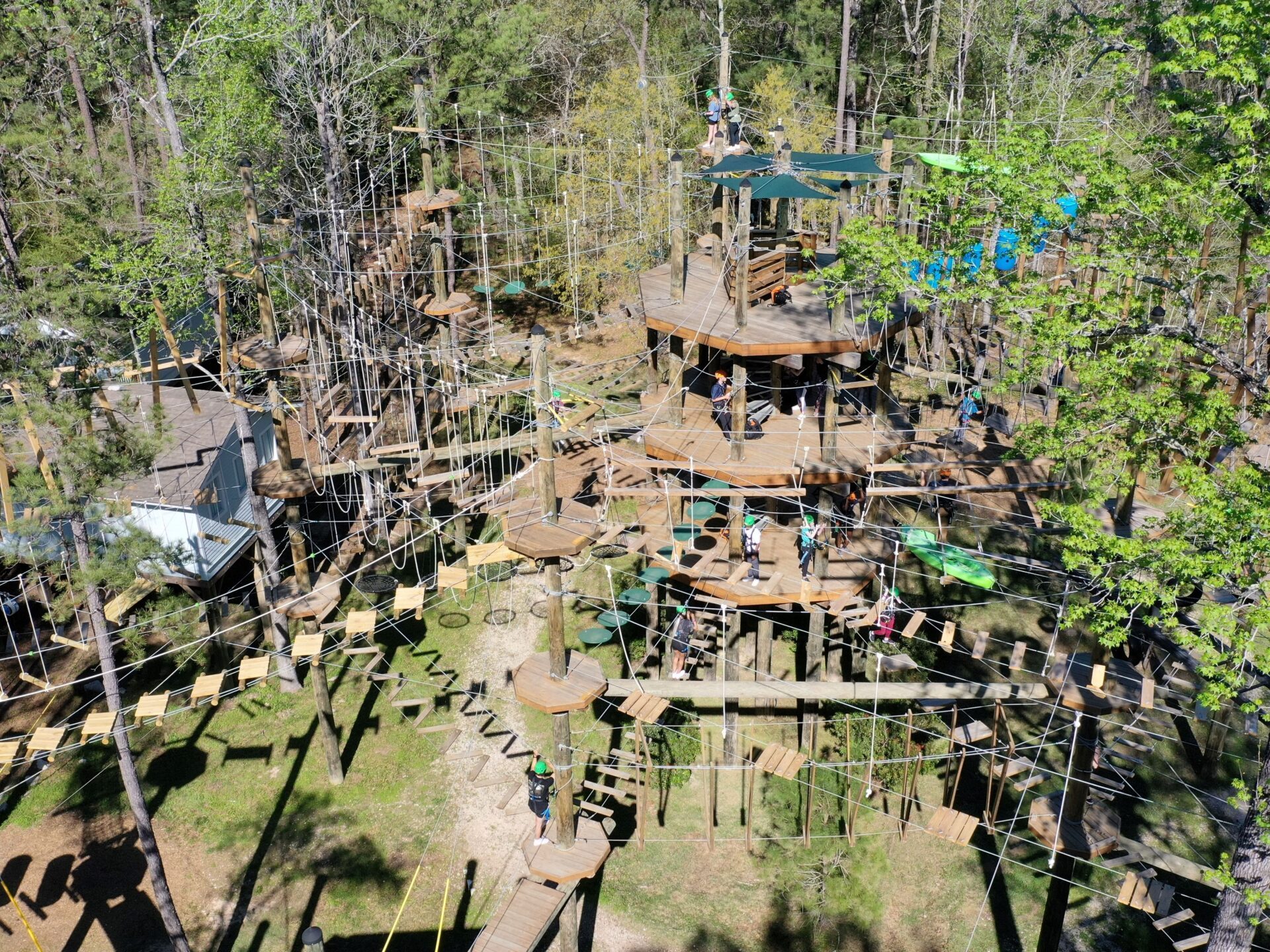 An aerial view of an extensive outdoor high ropes course set in a forested area. The course includes multiple levels of wooden platforms, rope bridges, and various obstacles. Participants wearing helmets and safety harnesses are visible navigating the course. The dense trees and green foliage indicate the course is in a natural setting.