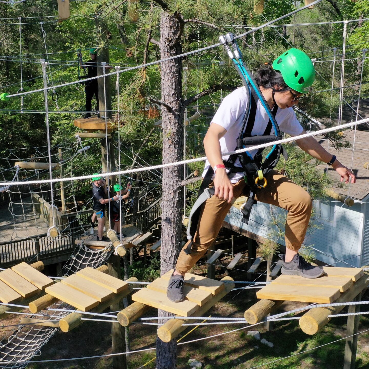 A group of people navigating a high ropes course in an outdoor setting. They are wearing green helmets and harnesses for safety. The participants are balancing on wooden planks suspended by ropes, with one person in the foreground carefully stepping forward. Trees and additional ropes course elements are visible in the background, along with a building partially seen through the trees.
