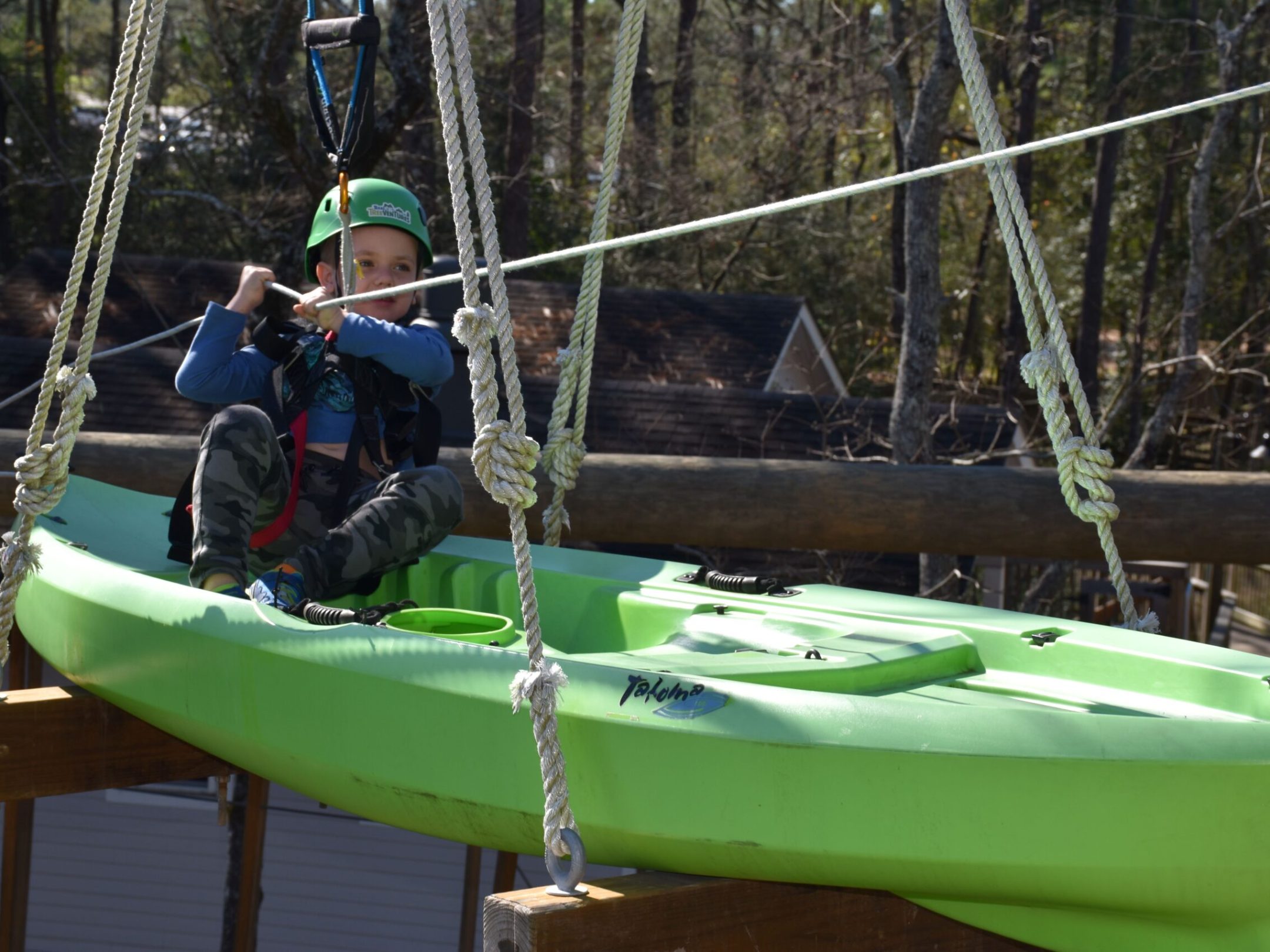 A child wearing a green helmet and safety harness sits in a green kayak suspended in the air as part of a high ropes course. The child is holding onto a rope for support. The background includes trees and rooftops, indicating an outdoor setting.