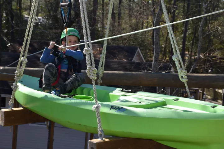 A child wearing a green helmet and safety harness sits in a green kayak suspended in the air as part of a high ropes course. The child is holding onto a rope for support. The background includes trees and rooftops, indicating an outdoor setting.
