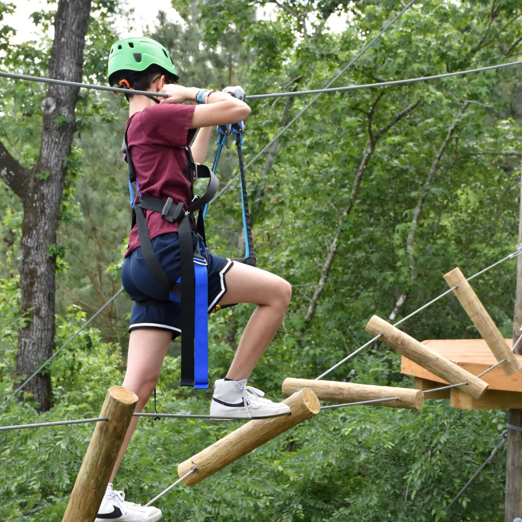 A person wearing a green helmet and safety harness navigates a high ropes course. They are stepping on irregularly spaced wooden logs suspended by ropes and holding onto higher ropes for balance. The person is dressed in a maroon T-shirt, blue shorts, and white sneakers. The background features lush green foliage, indicating an outdoor setting.