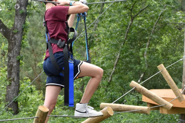 A person wearing a green helmet and safety harness navigates a high ropes course. They are stepping on irregularly spaced wooden logs suspended by ropes and holding onto higher ropes for balance. The person is dressed in a maroon T-shirt, blue shorts, and white sneakers. The background features lush green foliage, indicating an outdoor setting.