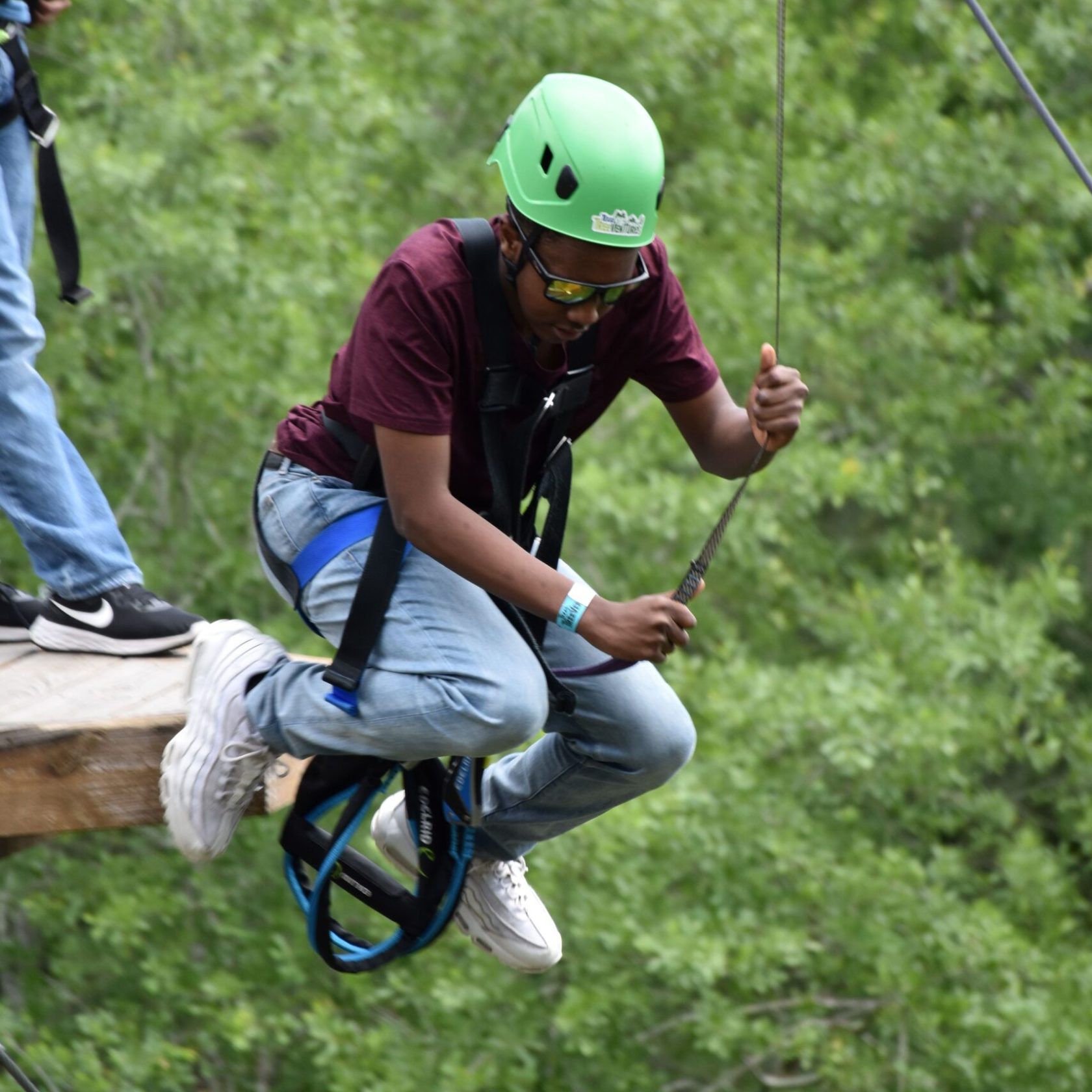 A person wearing a green helmet and safety harness prepares to step off a wooden platform on a high ropes course. They are holding onto a rope and dressed in a maroon T-shirt, light blue jeans, and white sneakers. The background features lush green foliage, indicating an outdoor setting.