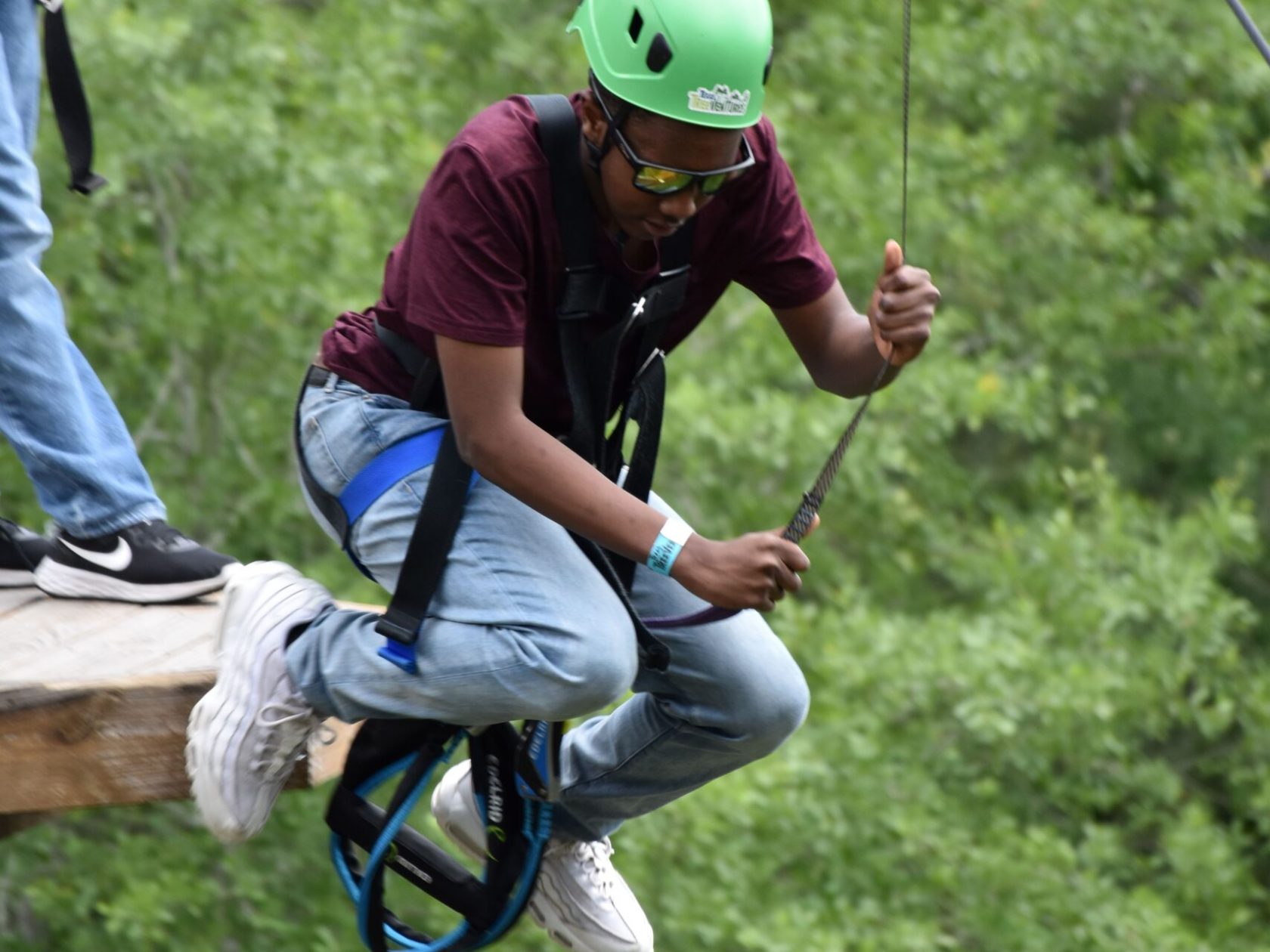A person wearing a green helmet and safety harness prepares to step off a wooden platform on a high ropes course. They are holding onto a rope and dressed in a maroon T-shirt, light blue jeans, and white sneakers. The background features lush green foliage, indicating an outdoor setting.