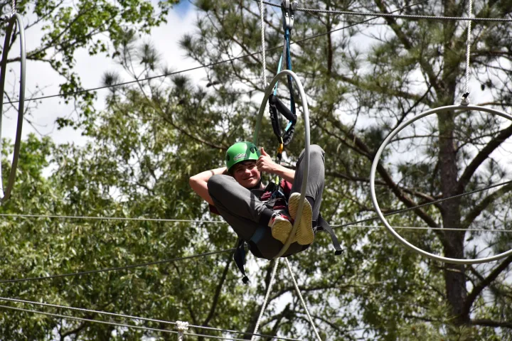 A person wearing a green helmet and safety harness is suspended in the air on a high ropes course. They are sitting inside a large metal ring and smiling, holding onto the ring for balance. The background features trees and a partly cloudy sky, indicating an outdoor setting.