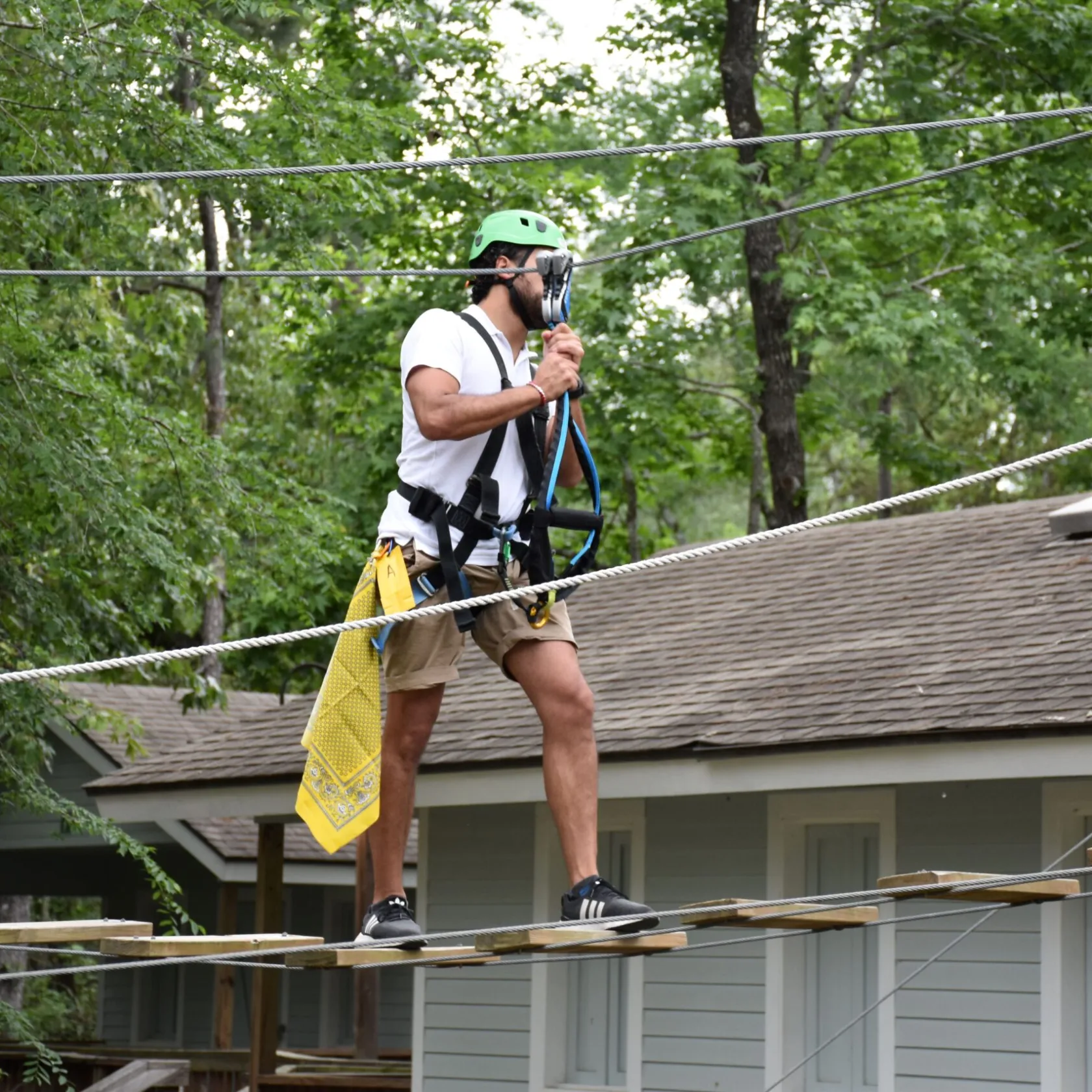 A man wearing a green helmet and safety harness navigates a high ropes course. He is walking on narrow wooden planks suspended by ropes and holding onto higher ropes for balance. The man is dressed in a white T-shirt, khaki shorts, and black sneakers, with a yellow cloth hanging from his belt. The background features trees and a building with a sloped roof, indicating an outdoor setting.