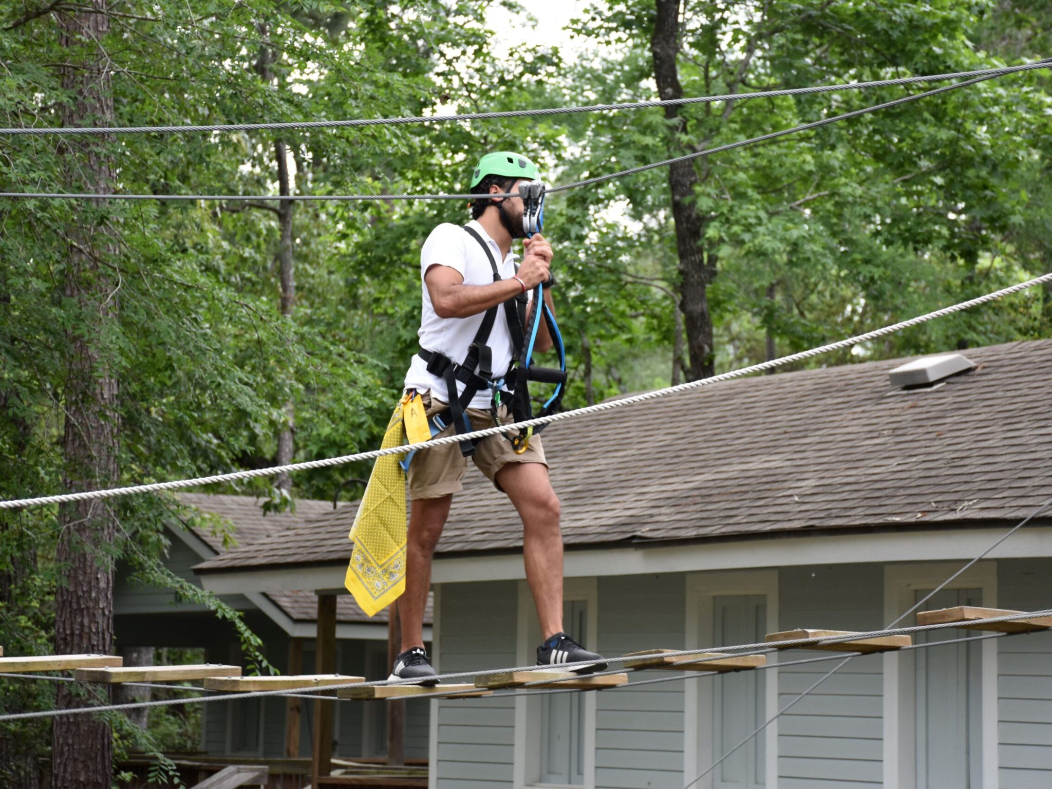 A man wearing a green helmet and safety harness navigates a high ropes course. He is walking on narrow wooden planks suspended by ropes and holding onto higher ropes for balance. The man is dressed in a white T-shirt, khaki shorts, and black sneakers, with a yellow cloth hanging from his belt. The background features trees and a building with a sloped roof, indicating an outdoor setting.