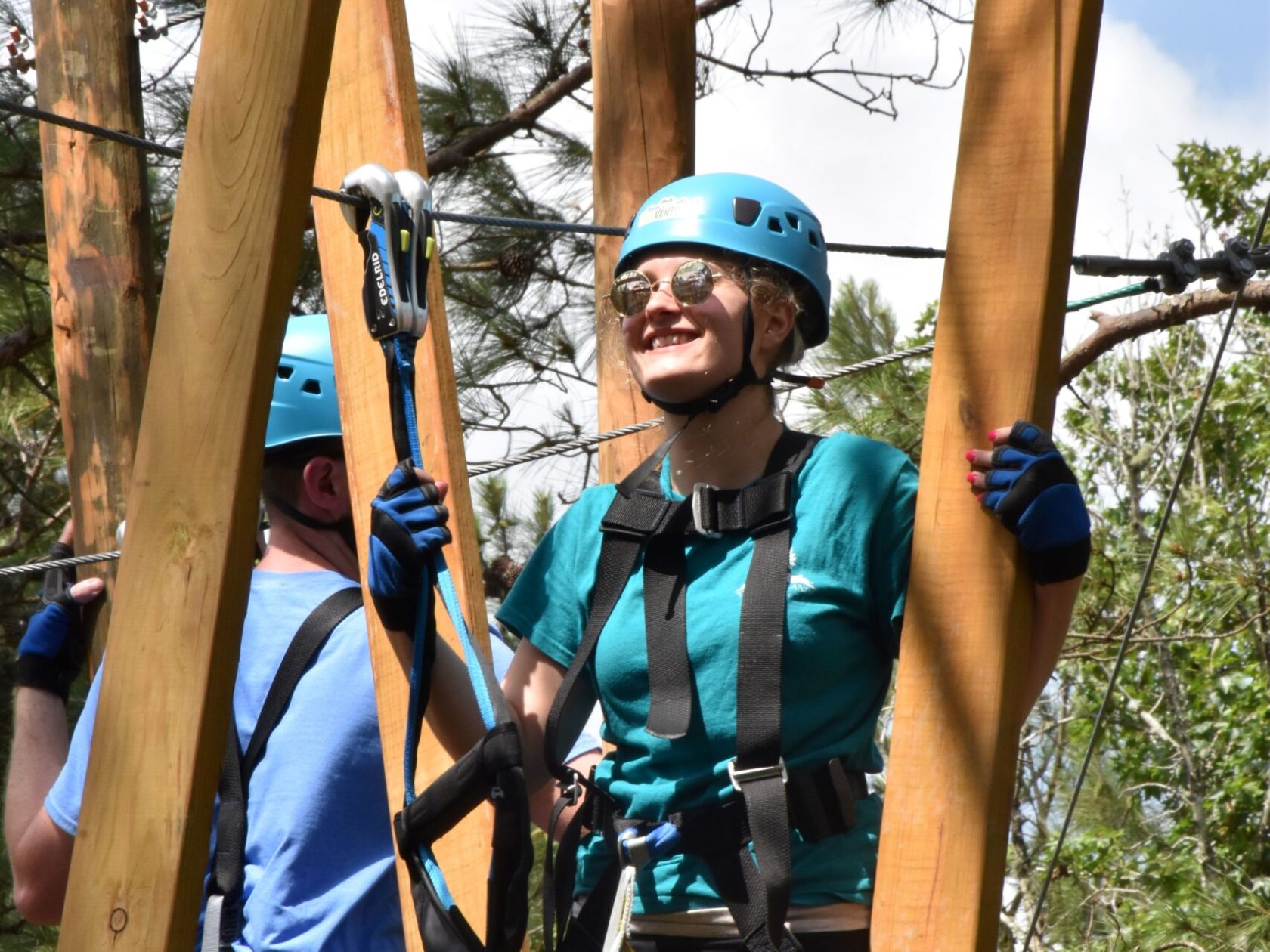 A woman wearing a blue helmet and safety harness smiles as she navigates a high ropes course. She is holding onto wooden beams and wearing a turquoise T-shirt, black leggings, and gloves. Another person in a blue shirt and helmet is partially visible in the background. The setting includes trees and a clear sky, indicating an outdoor environment.