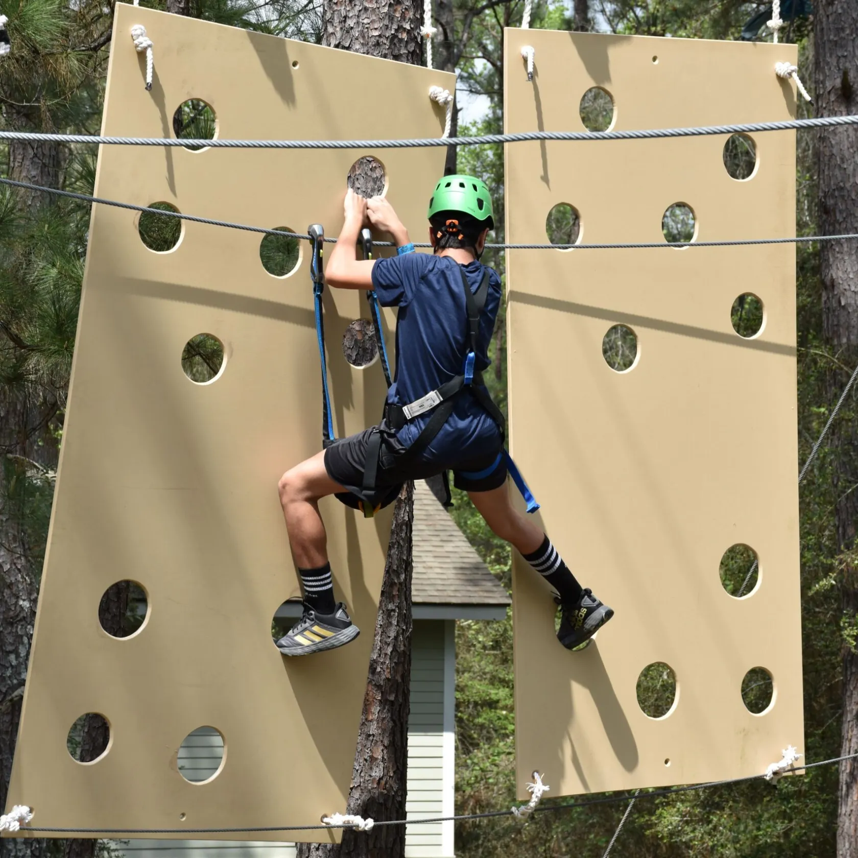 A person wearing a green helmet and safety harness climbs a vertical obstacle on a high ropes course. The obstacle consists of large beige panels with circular holes. The person is dressed in a blue shirt, black shorts, and striped socks. The background features trees and foliage, indicating the course is set in a forested area.