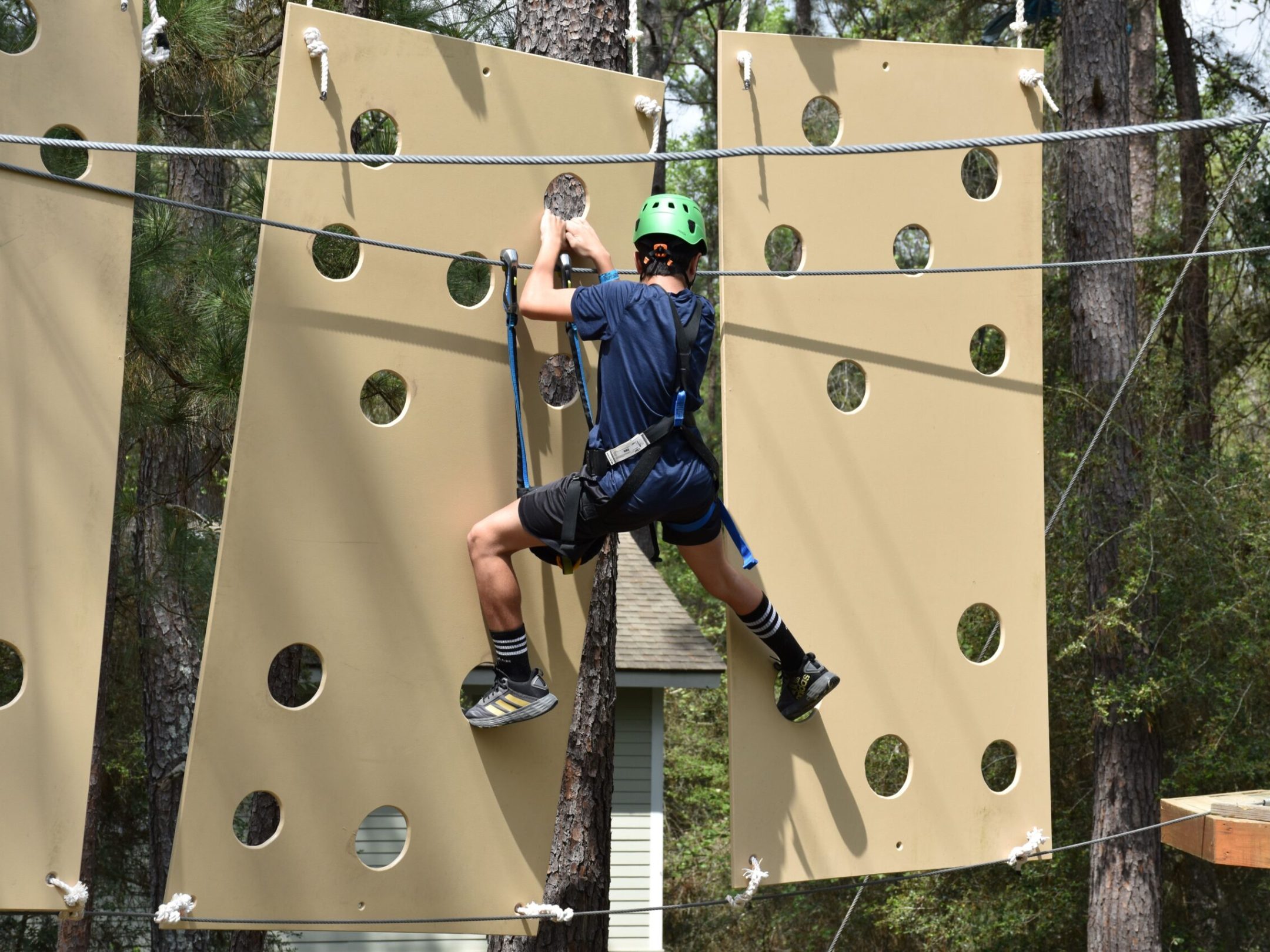 A person wearing a green helmet and safety harness climbs a vertical obstacle on a high ropes course. The obstacle consists of large beige panels with circular holes. The person is dressed in a blue shirt, black shorts, and striped socks. The background features trees and foliage, indicating the course is set in a forested area.