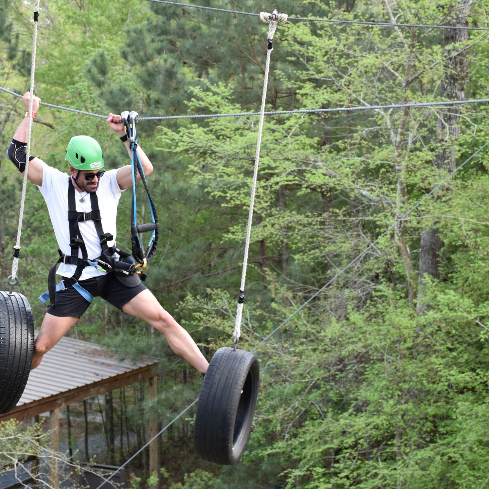 A man wearing a green helmet and safety harness navigates a high ropes course. He is stepping on hanging tires suspended by ropes, holding onto the ropes for balance. The man is dressed in a white T-shirt, black shorts, and sunglasses. The background features trees and foliage, indicating the course is set in a forested area.