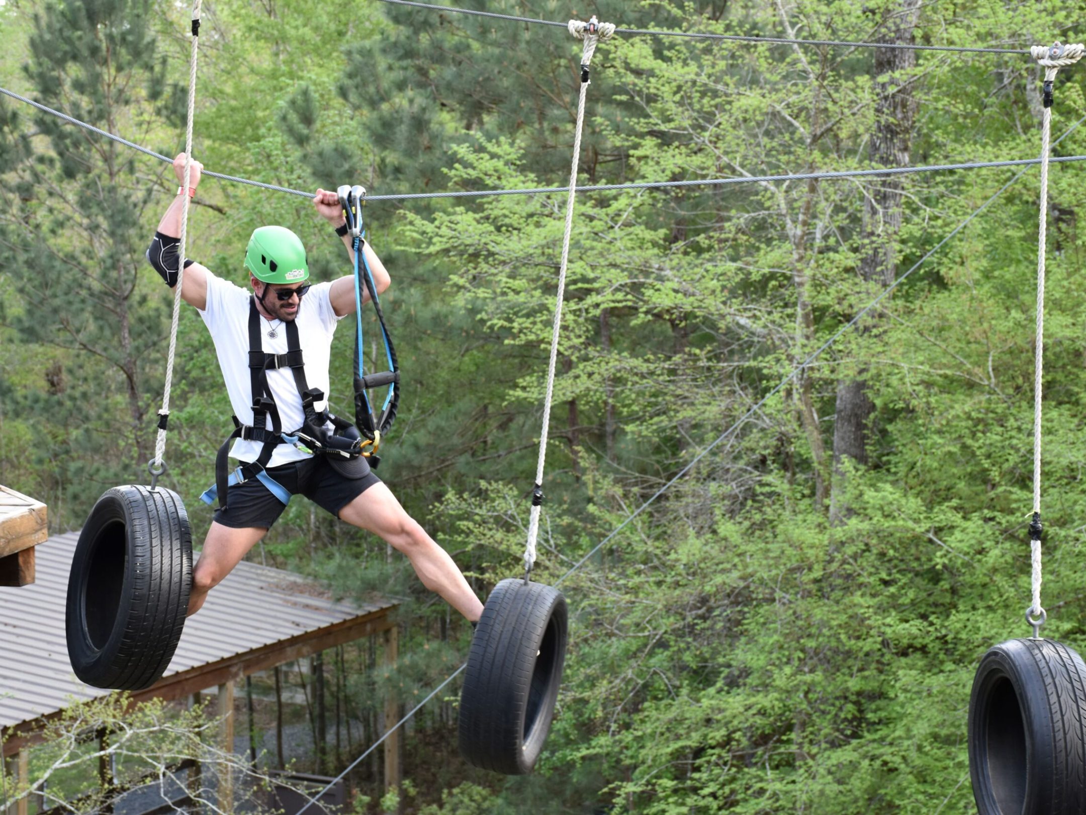 A man wearing a green helmet and safety harness navigates a high ropes course. He is stepping on hanging tires suspended by ropes, holding onto the ropes for balance. The man is dressed in a white T-shirt, black shorts, and sunglasses. The background features trees and foliage, indicating the course is set in a forested area.