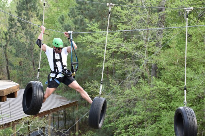A man wearing a green helmet and safety harness navigates a high ropes course. He is stepping on hanging tires suspended by ropes, holding onto the ropes for balance. The man is dressed in a white T-shirt, black shorts, and sunglasses. The background features trees and foliage, indicating the course is set in a forested area.