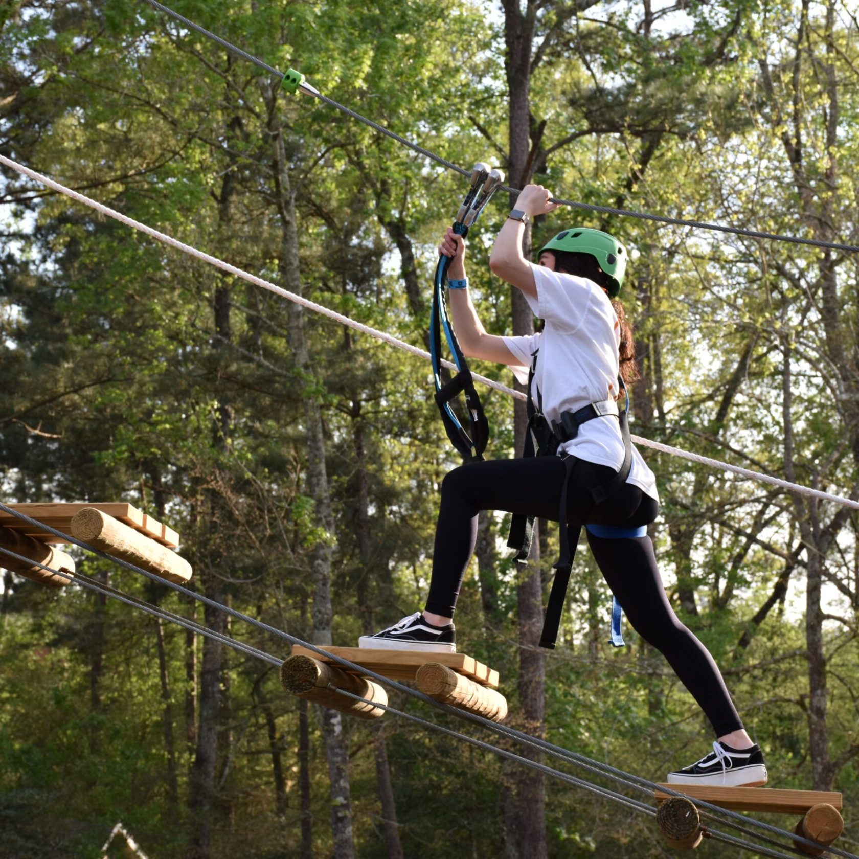 A person wearing a green helmet and safety harness navigates a high ropes course. They are stepping on narrow wooden planks suspended by ropes and holding onto higher ropes for balance. The person is dressed in a white T-shirt, black leggings, and sneakers. The background features trees and foliage, indicating the course is set in a forested area.