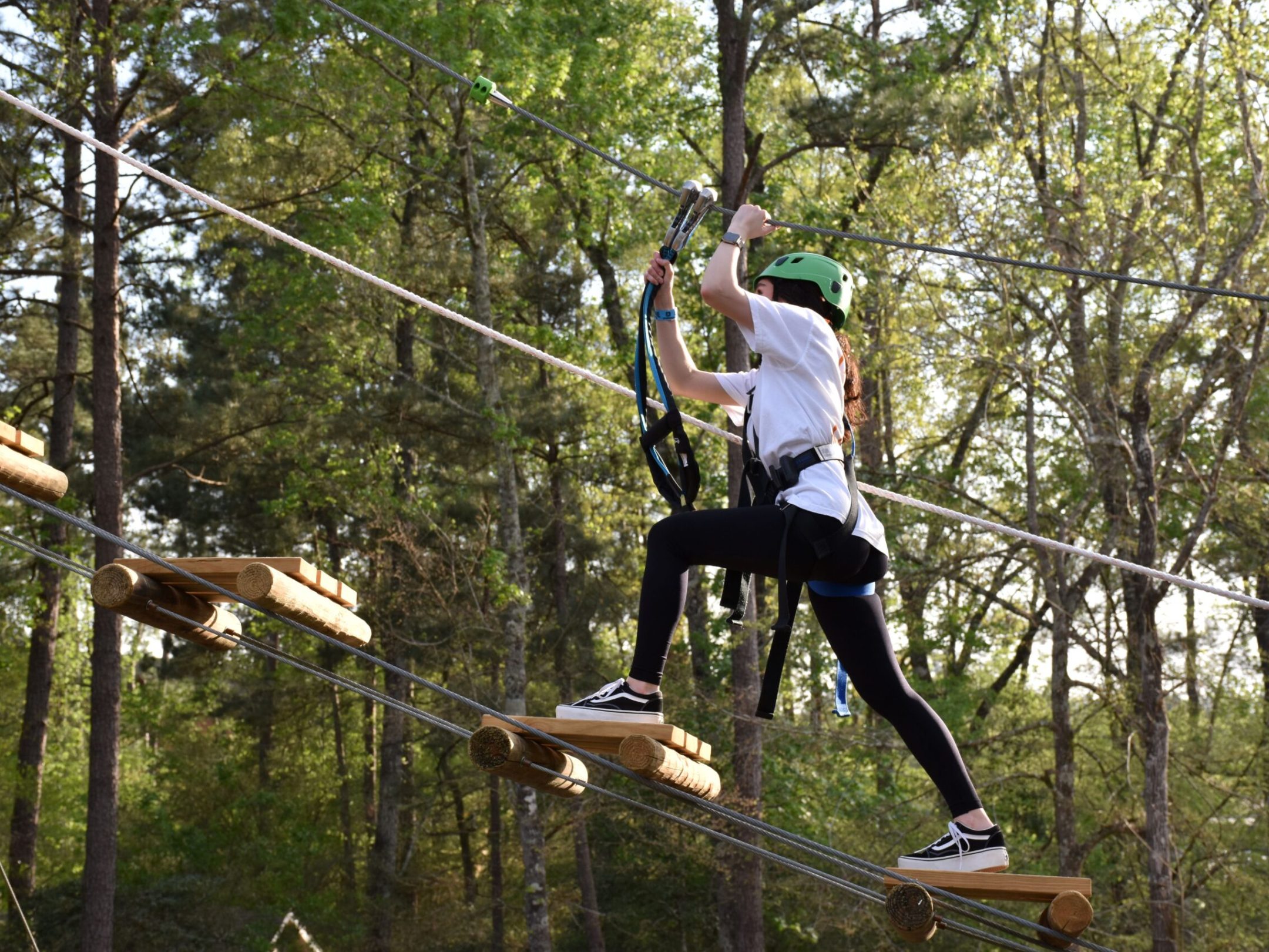 A person wearing a green helmet and safety harness navigates a high ropes course. They are stepping on narrow wooden planks suspended by ropes and holding onto higher ropes for balance. The person is dressed in a white T-shirt, black leggings, and sneakers. The background features trees and foliage, indicating the course is set in a forested area.