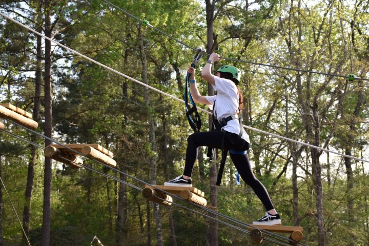 A person wearing a green helmet and safety harness navigates a high ropes course. They are stepping on narrow wooden planks suspended by ropes and holding onto higher ropes for balance. The person is dressed in a white T-shirt, black leggings, and sneakers. The background features trees and foliage, indicating the course is set in a forested area.