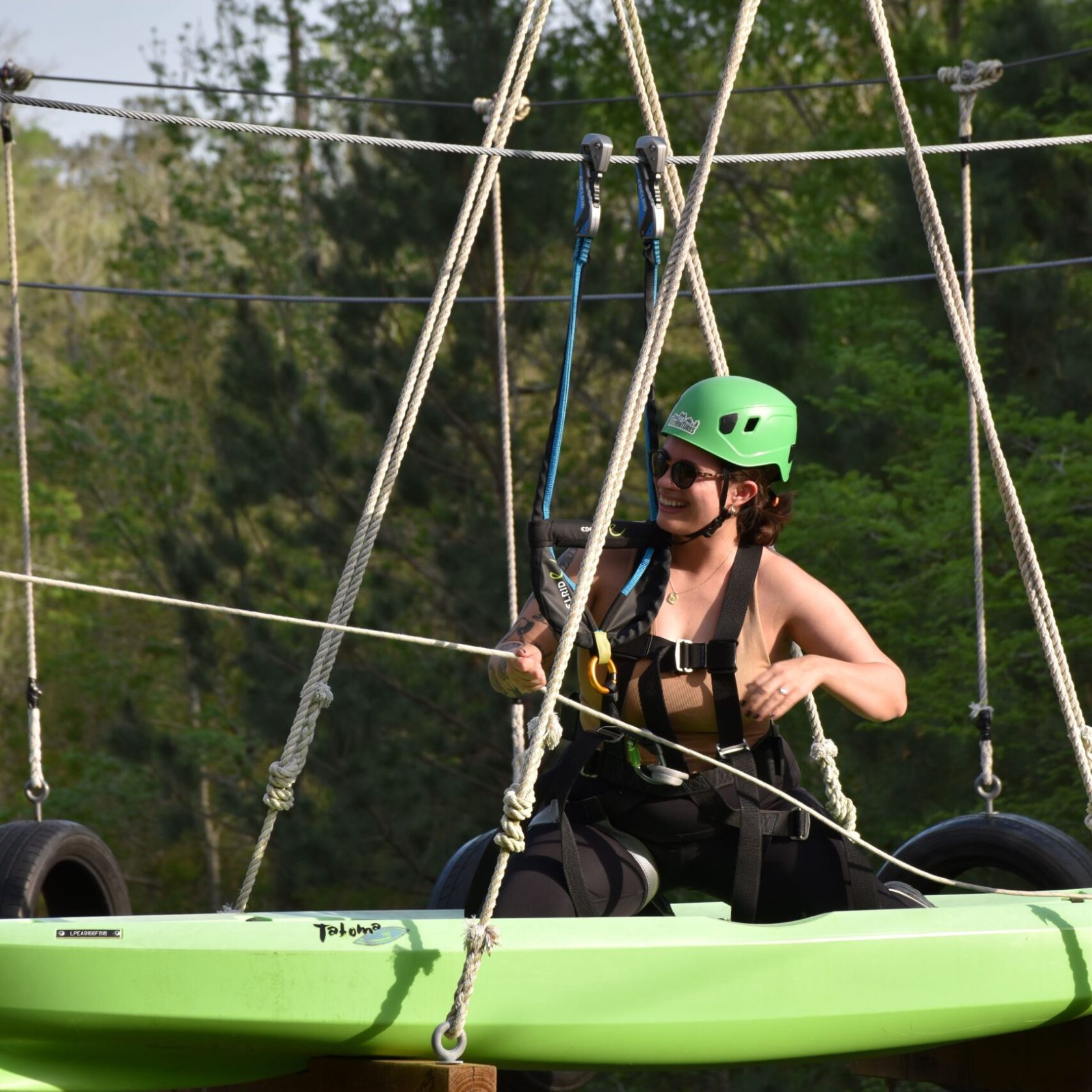 A person wearing a green helmet and safety harness is sitting in a green kayak suspended in the air. They are holding onto ropes with a smile on their face, wearing sunglasses and a sleeveless black top. The kayak is part of a high ropes course, with tires and ropes visible in the background. The setting is outdoors, with lush green trees in the background.