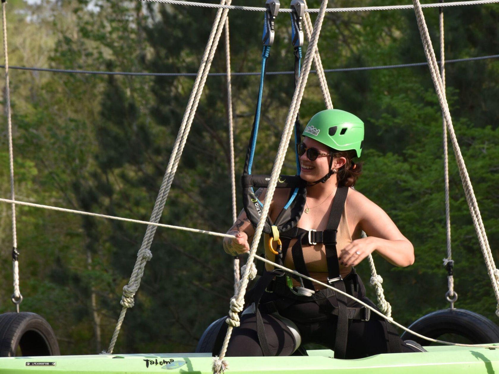 A person wearing a green helmet and safety harness is sitting in a green kayak suspended in the air. They are holding onto ropes with a smile on their face, wearing sunglasses and a sleeveless black top. The kayak is part of a high ropes course, with tires and ropes visible in the background. The setting is outdoors, with lush green trees in the background.