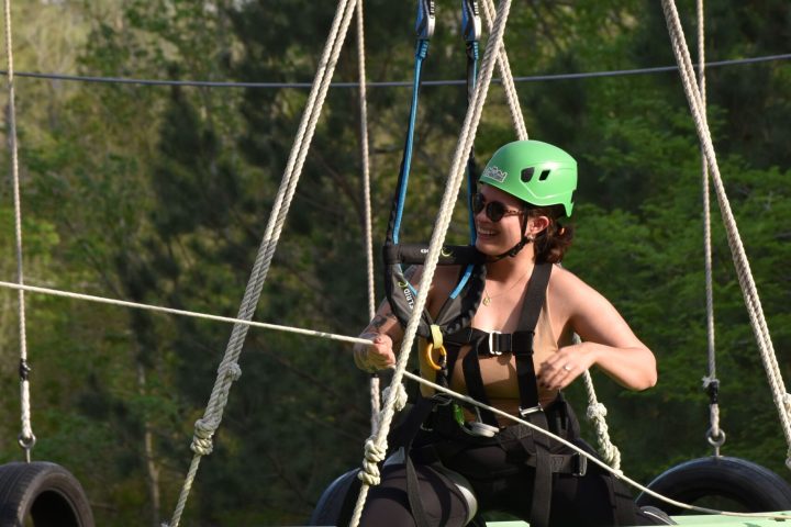 A person wearing a green helmet and safety harness is sitting in a green kayak suspended in the air. They are holding onto ropes with a smile on their face, wearing sunglasses and a sleeveless black top. The kayak is part of a high ropes course, with tires and ropes visible in the background. The setting is outdoors, with lush green trees in the background.
