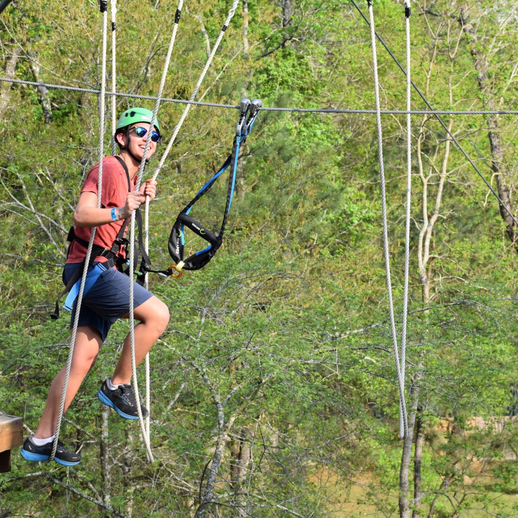 A person wearing a green helmet and safety harness is navigating a high ropes course. They are holding onto ropes and are suspended in the air, with a platform visible to the left. The person is smiling and wearing sunglasses, a red T-shirt, blue shorts, and athletic shoes. The background features lush green trees, indicating an outdoor setting.