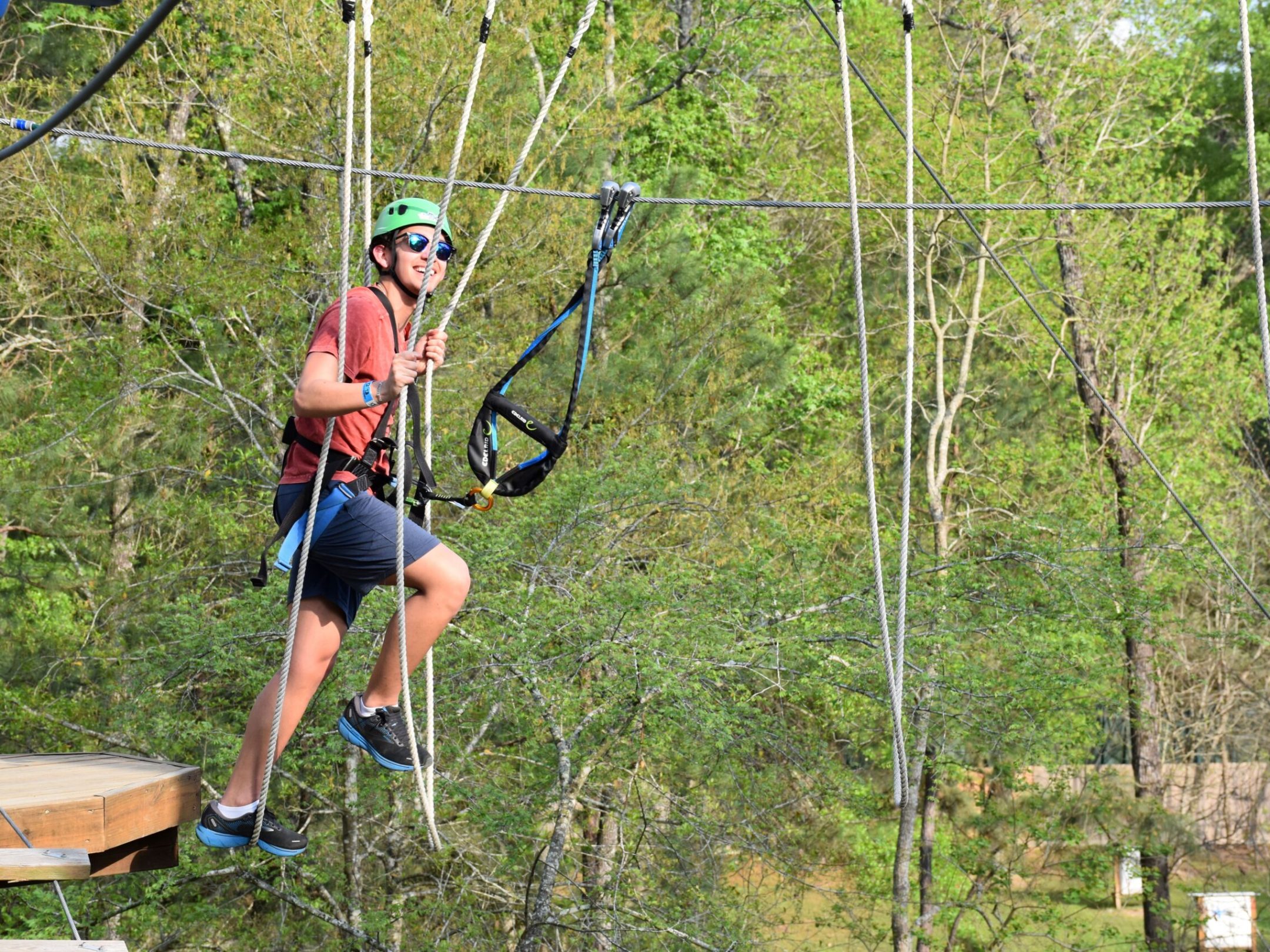 A person wearing a green helmet and safety harness is navigating a high ropes course. They are holding onto ropes and are suspended in the air, with a platform visible to the left. The person is smiling and wearing sunglasses, a red T-shirt, blue shorts, and athletic shoes. The background features lush green trees, indicating an outdoor setting.
