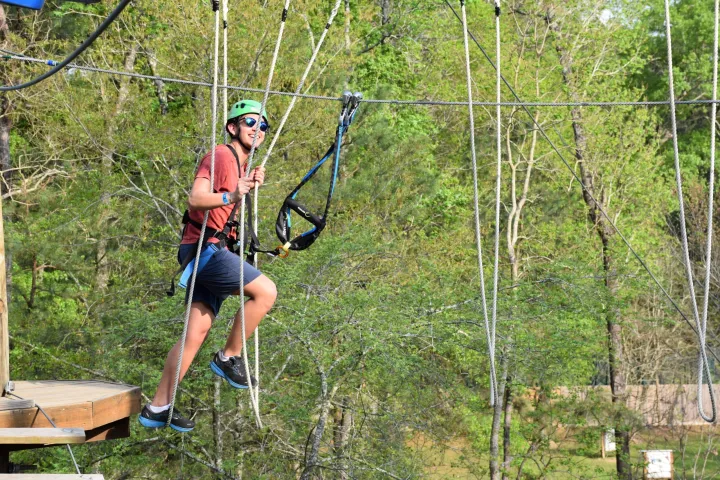 A person wearing a green helmet and safety harness is navigating a high ropes course. They are holding onto ropes and are suspended in the air, with a platform visible to the left. The person is smiling and wearing sunglasses, a red T-shirt, blue shorts, and athletic shoes. The background features lush green trees, indicating an outdoor setting.